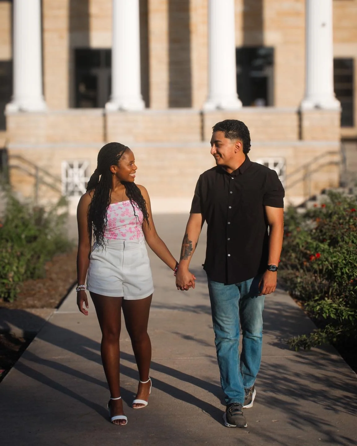 A young couple walking hand-in-hand outside, smiling at each other, with a building and some greenery in the background.