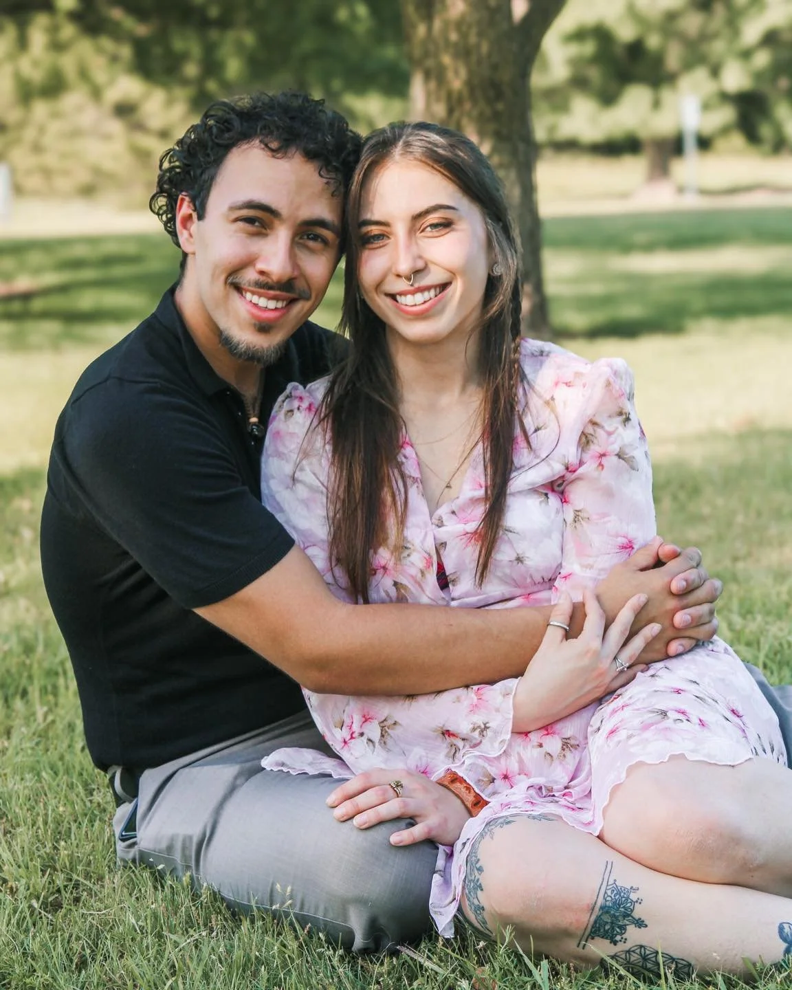 A couple sitting on the grass in a park, smiling, with trees in the background.
