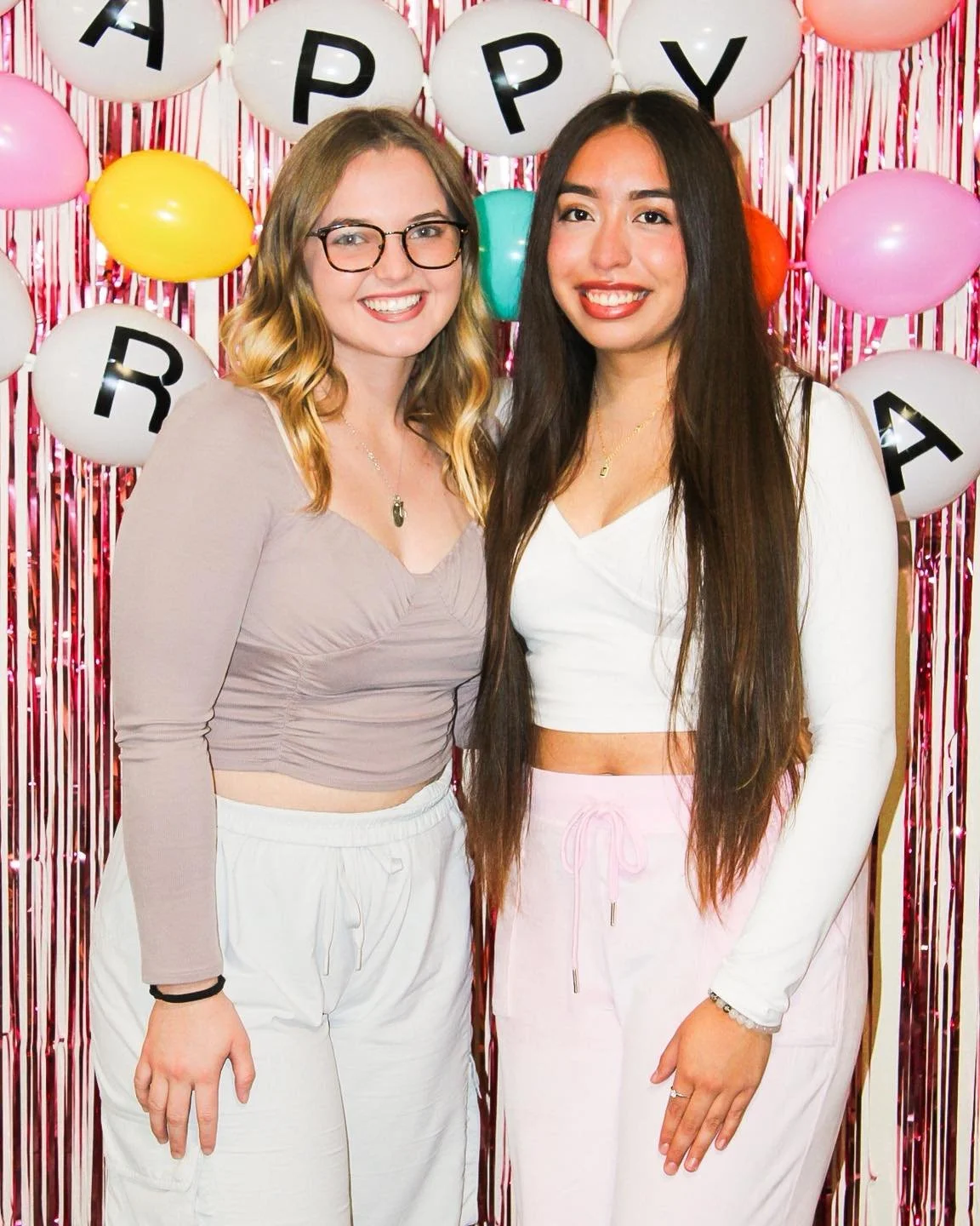 Two women standing in front of a colorful birthday party backdrop with pink, white, yellow, green, and orange balloons, and black and white balloons spelling 'HAPPY BIRTHDAY'. They are smiling and posing for the photo.