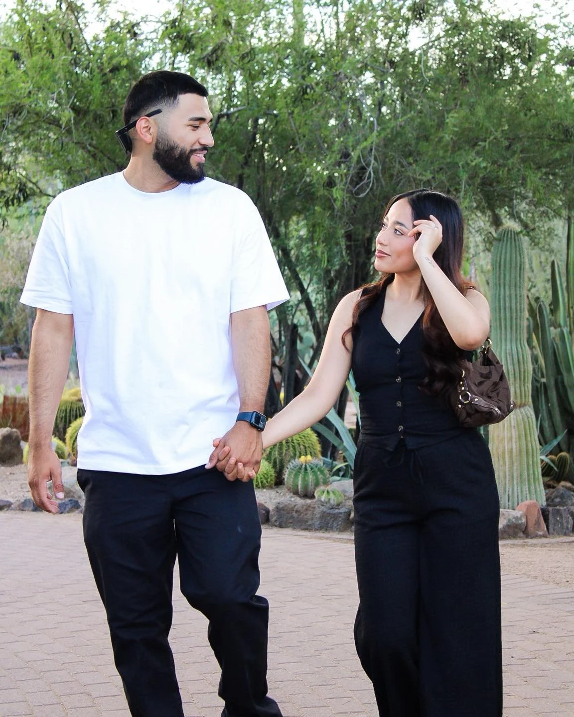 A man and woman walking hand-in-hand outdoors among cactus and plants, smiling at each other.