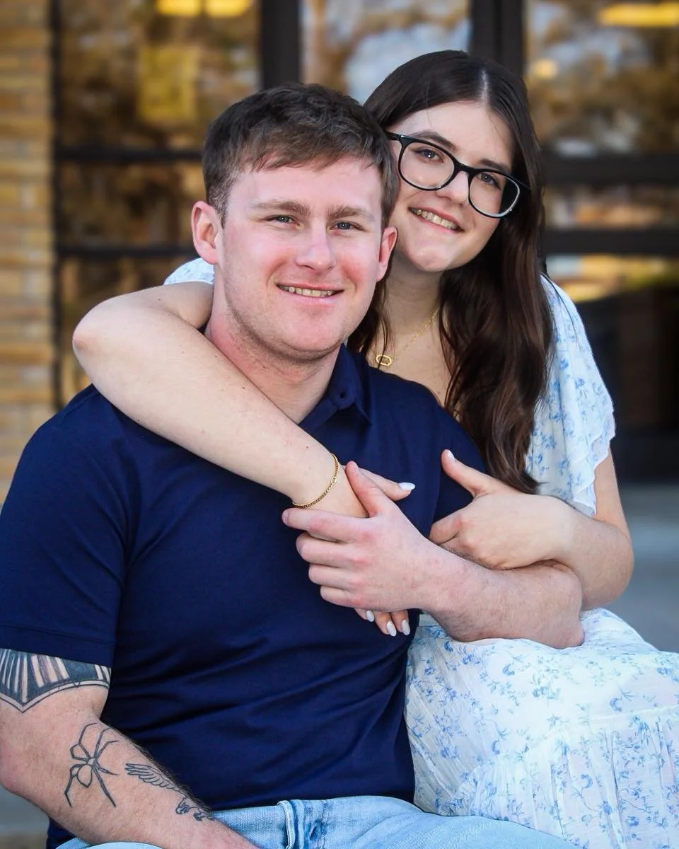 A young couple is smiling, with the woman embracing the man from behind. They are outdoors, with a building and some trees in the background.