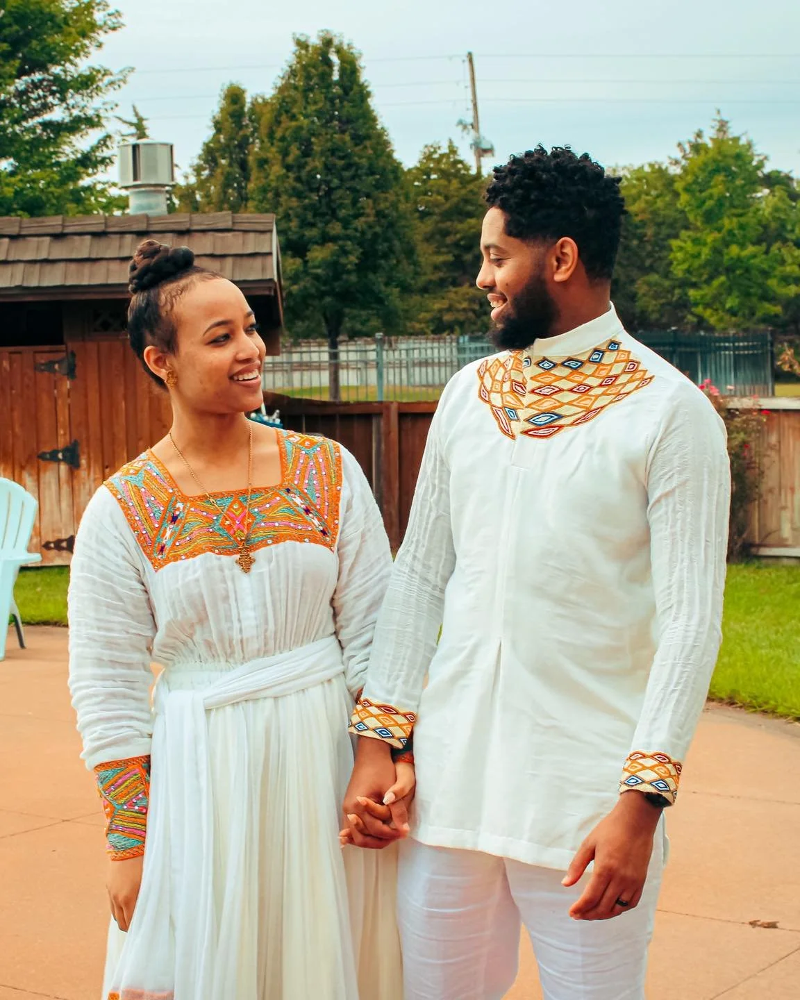 A couple dressed in traditional white clothing with colorful embroidered details holding hands and smiling at each other outdoors in a backyard.