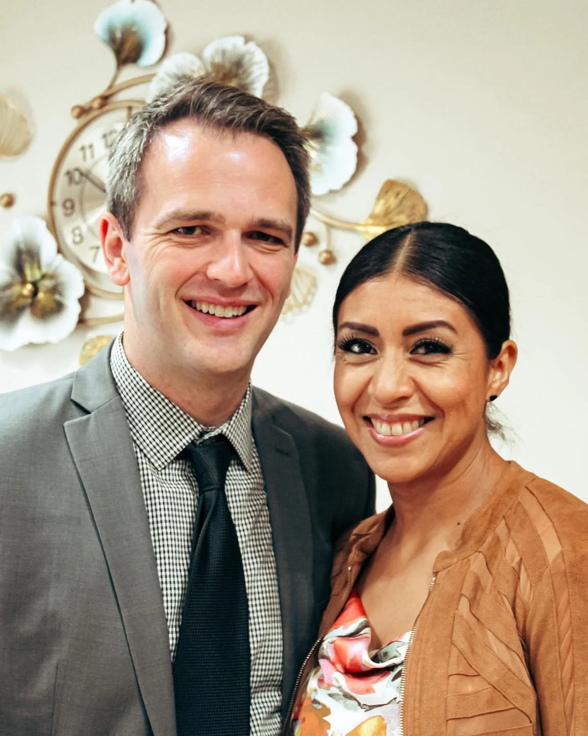 A smiling man in a gray suit and tie standing next to a smiling woman in a brown jacket and floral top, with decorative wall art and a clock in the background.