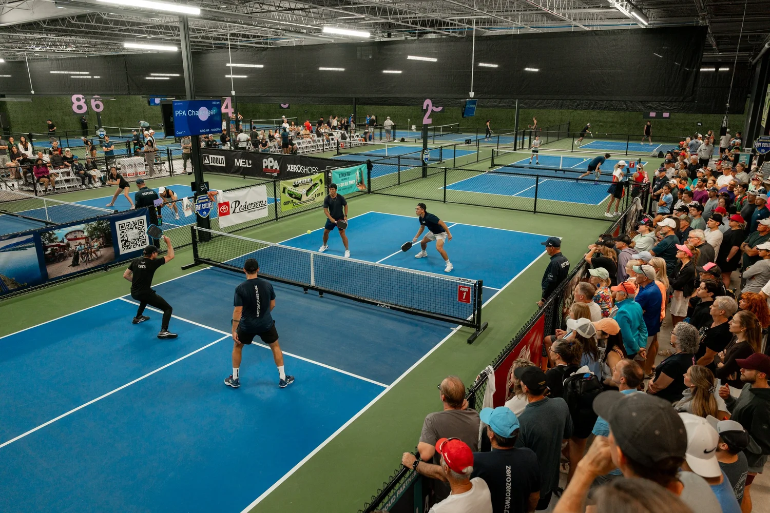 Doubles play on the indoor courts at Zero Zero Two, with spectators along the fence.