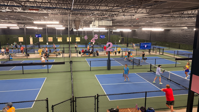 Indoor pickleball courts with players and officials, blue and green court surfaces, net barriers, and lighting fixtures overhead.