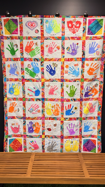 Colorful quilt with squares featuring handprint art in various colors and patterns, bordered with a multicolored patchwork design.