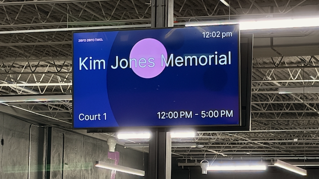 Electronic display board showing information for Kim Jones Memorial, located on Court 1, from 12:00 PM to 5:00 PM, with the current time 12:02 PM, at an indoor sports facility.