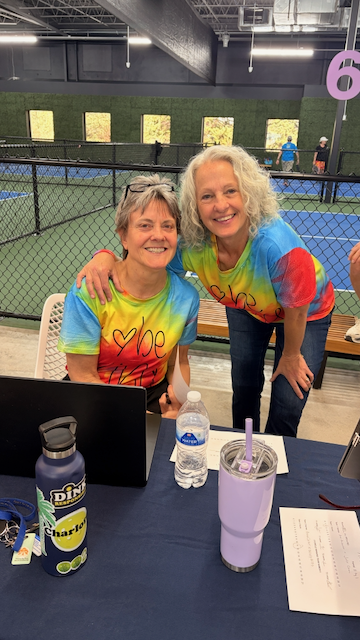 Two smiling women wearing matching multicolored tie-dye shirts with hearts and text, sitting at a table near an indoor tennis court, with water bottles and a laptop in front of them.