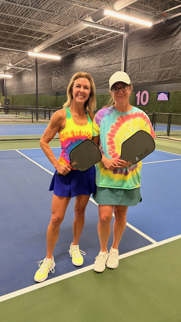 Two women standing on an indoor pickleball court, holding paddles and smiling. They are wearing colorful tie-dye shirts and skirts, with one wearing a white cap. The court has blue and green flooring, and there are tennis nets and a scoreboard in the