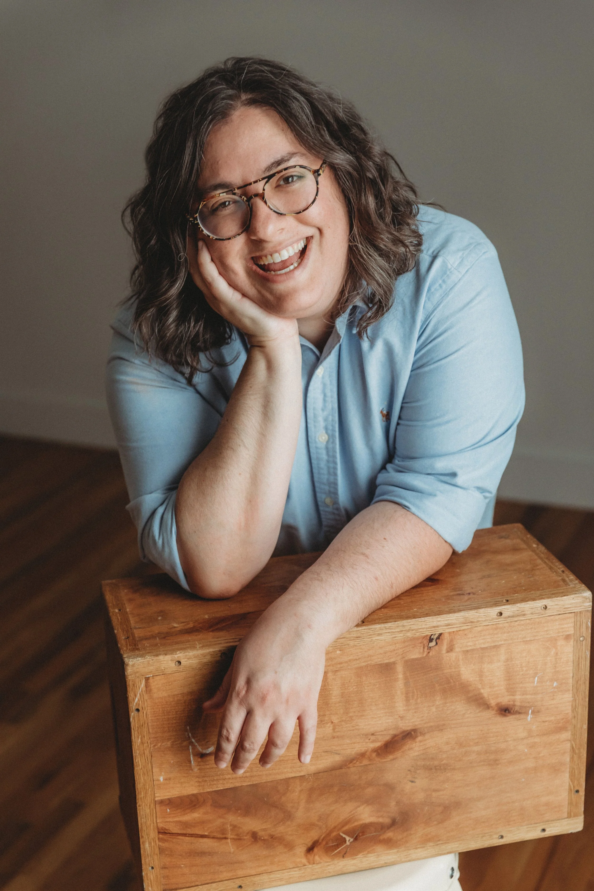 Smiling person with curly hair and glasses leaning on a wooden box.