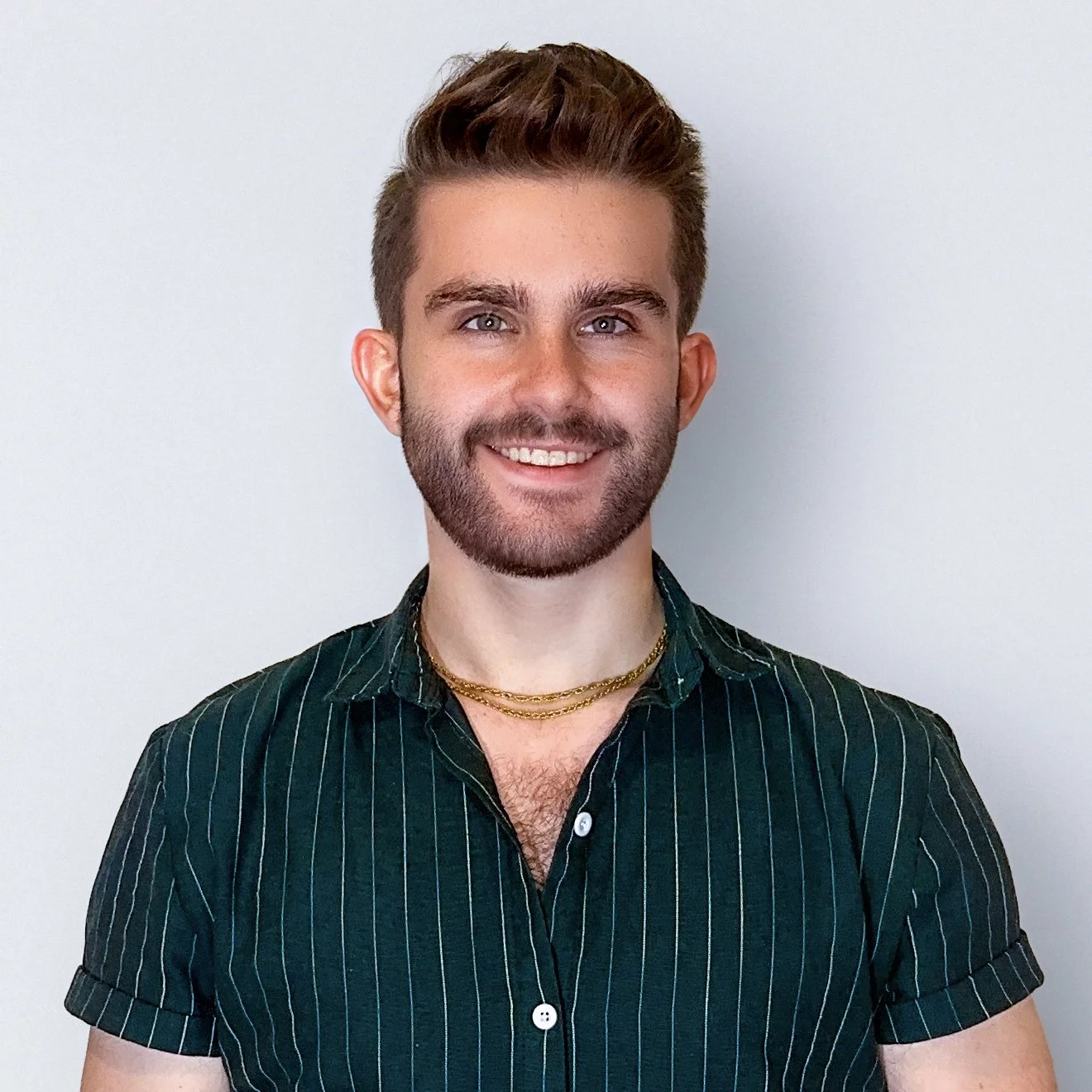 Adam Pozdro. A young man with styled brown hair, blue eyes, and a beard, smiling at the camera, wearing a dark green striped button-up shirt with the top few buttons open and layered gold necklaces against a plain light-colored background.