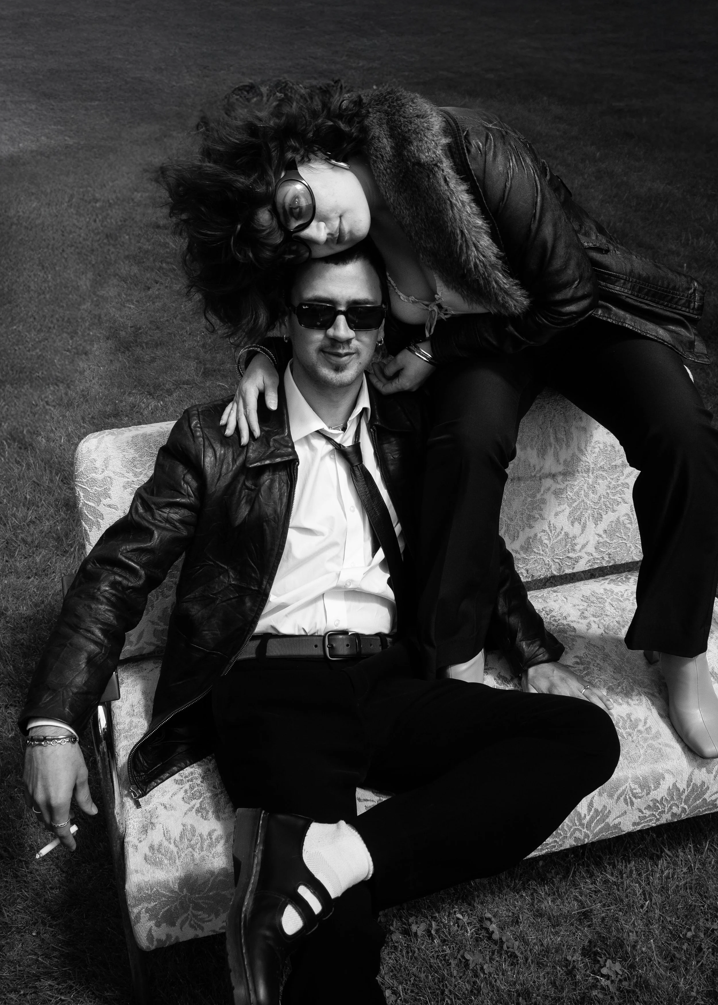Two stylish individuals, one sitting on a patterned sofa and the other leaning over, both wearing sunglasses and leather jackets, posed together outdoors on grass.