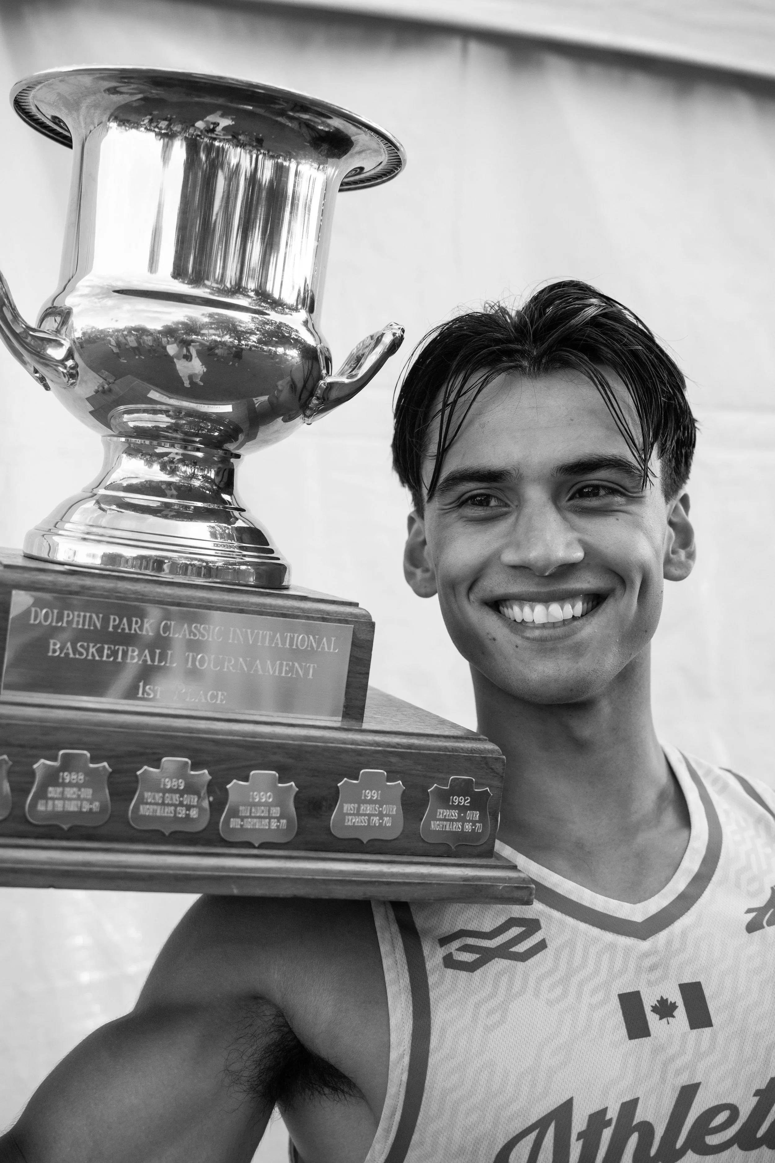 A young man smiling and holding a large trophy with a plaque that reads 'Dolphin Park Classic Invitational Basketball Tournament 1st Place.' He is wearing a sports jersey with a maple leaf and the word 'Athletic' printed on it.