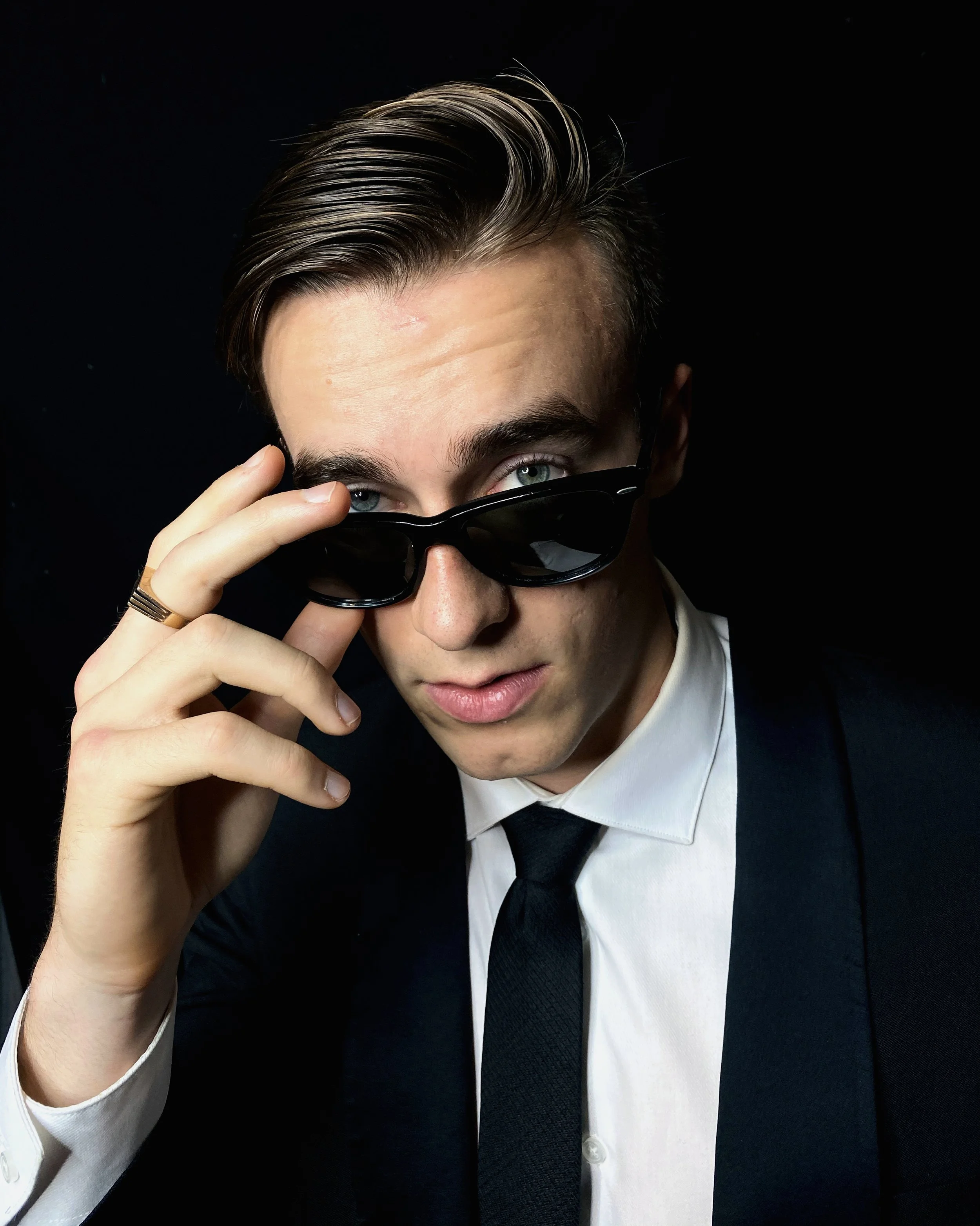 Vancouver BC professional headshot photography. A young man with slicked-back hair wearing sunglasses, a white shirt, a black tie, and a black suit against a black background.