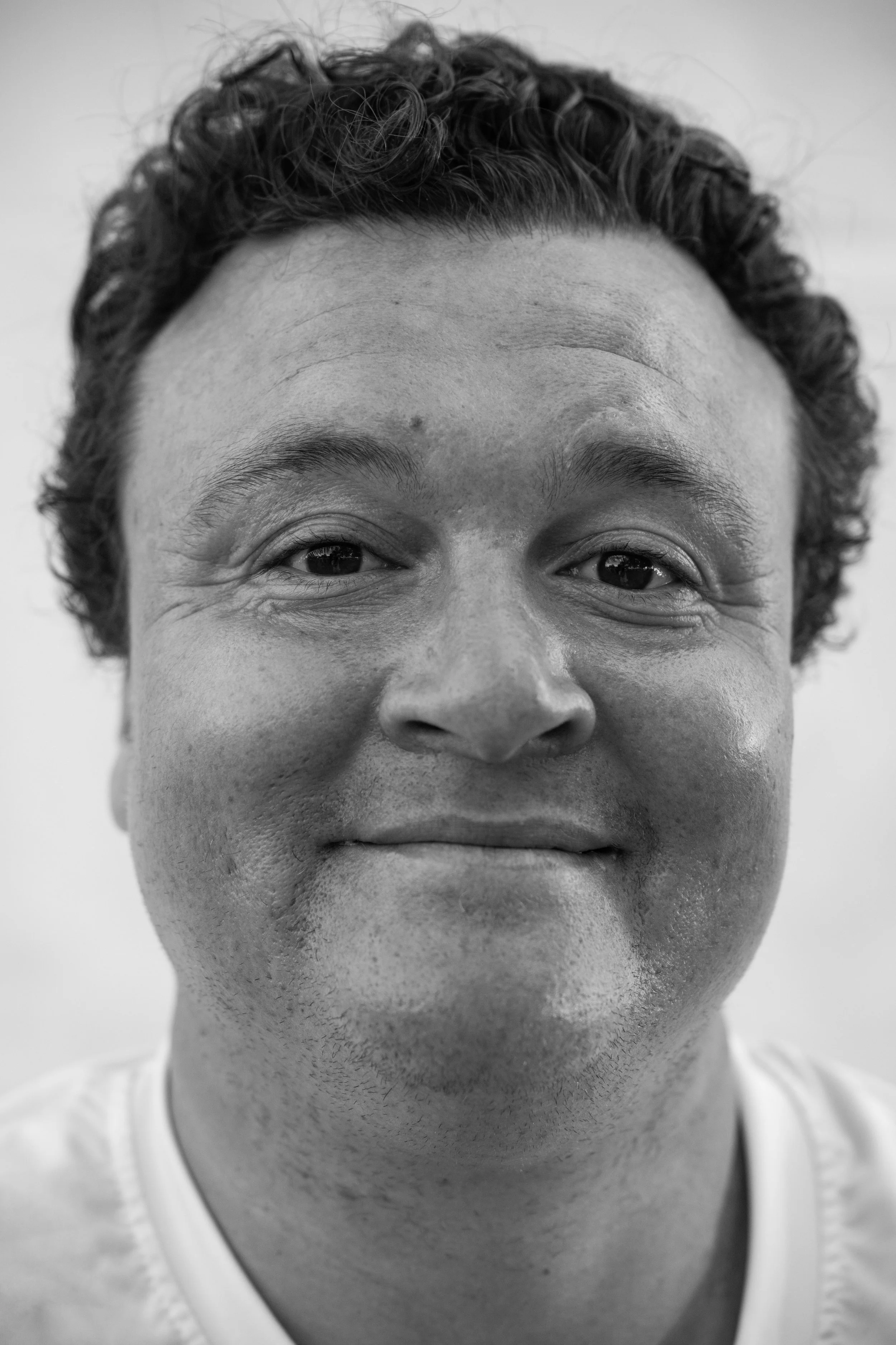 Black and white close-up portrait of a man with curly hair, smiling gently, wearing a white shirt.