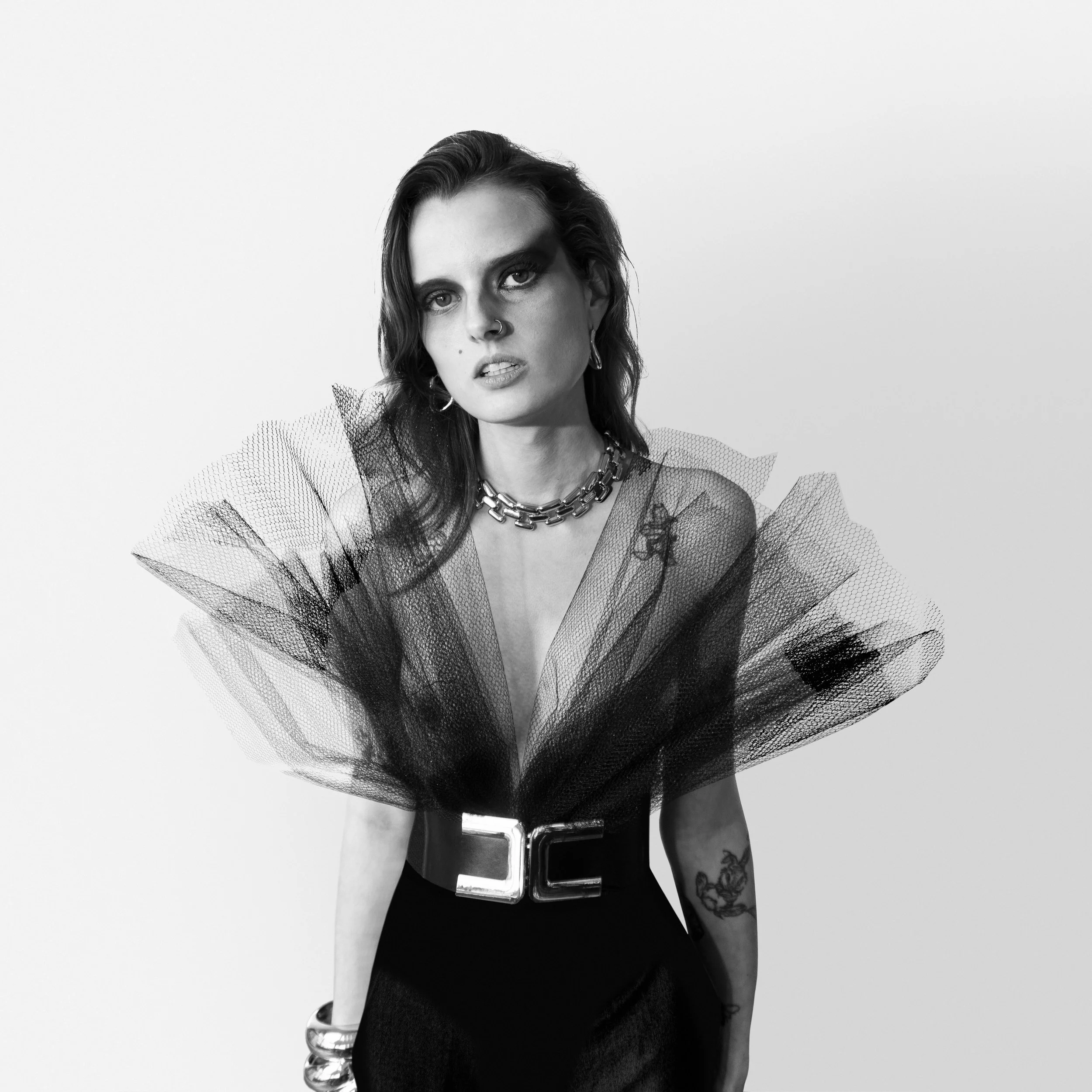 Vancouver BC professional headshot photography. Black and white portrait of a woman with dark hair, wearing a bold necklace, earrings, and a tulle dress with large ruffled shoulders and a belt, standing against a plain background.
