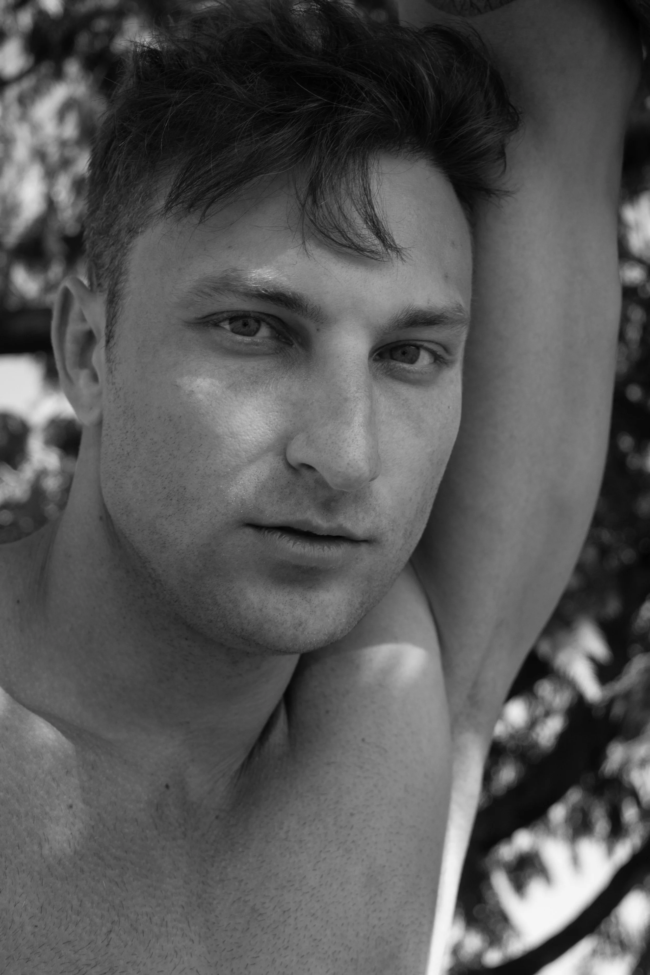 A black and white close-up portrait of a young man with short, tousled hair, leaning against a tree with one arm raised and resting on his head, outdoors with blurred foliage in the background.