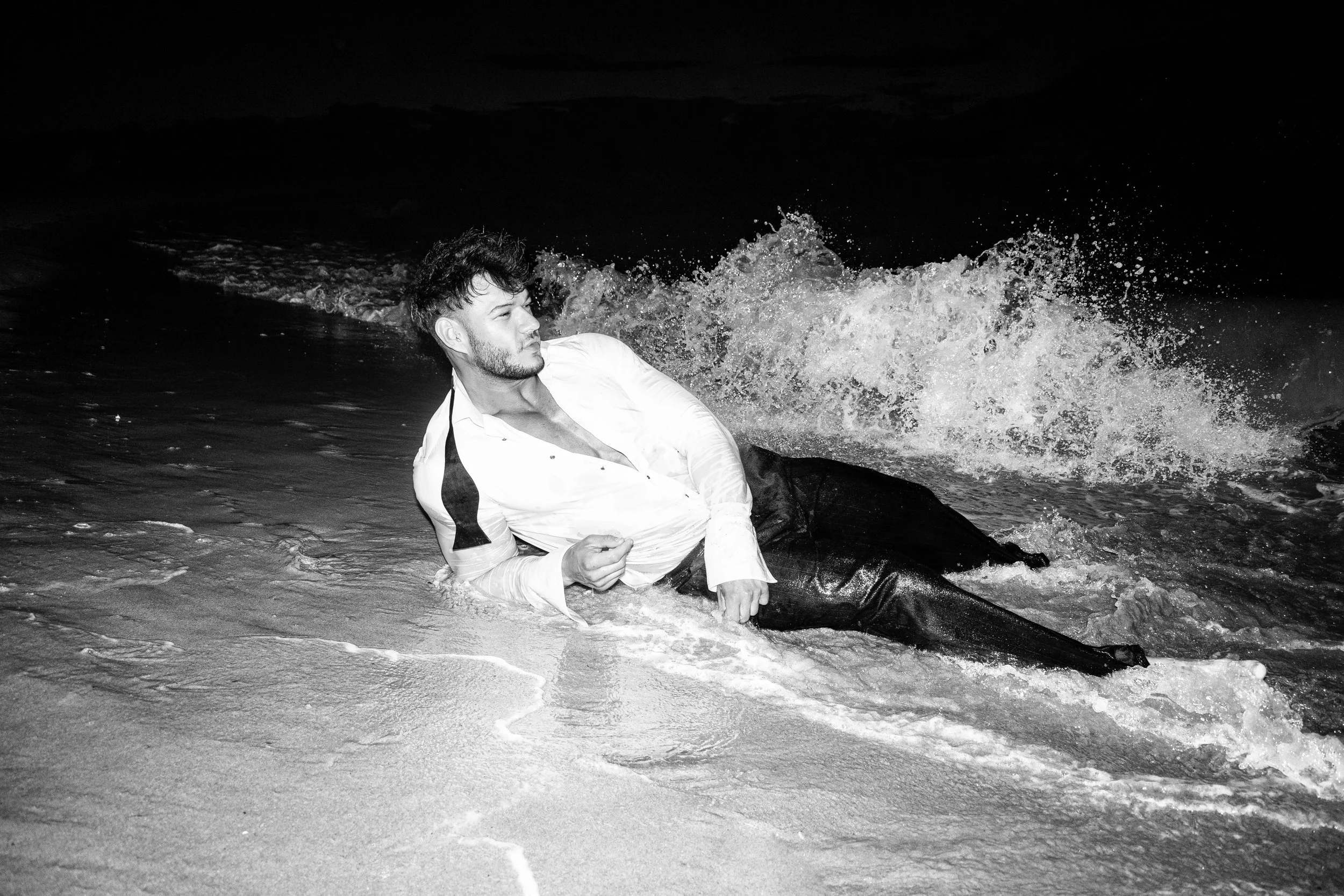 A man in a white shirt and dark pants lying on the beach with waves crashing around him at night.
