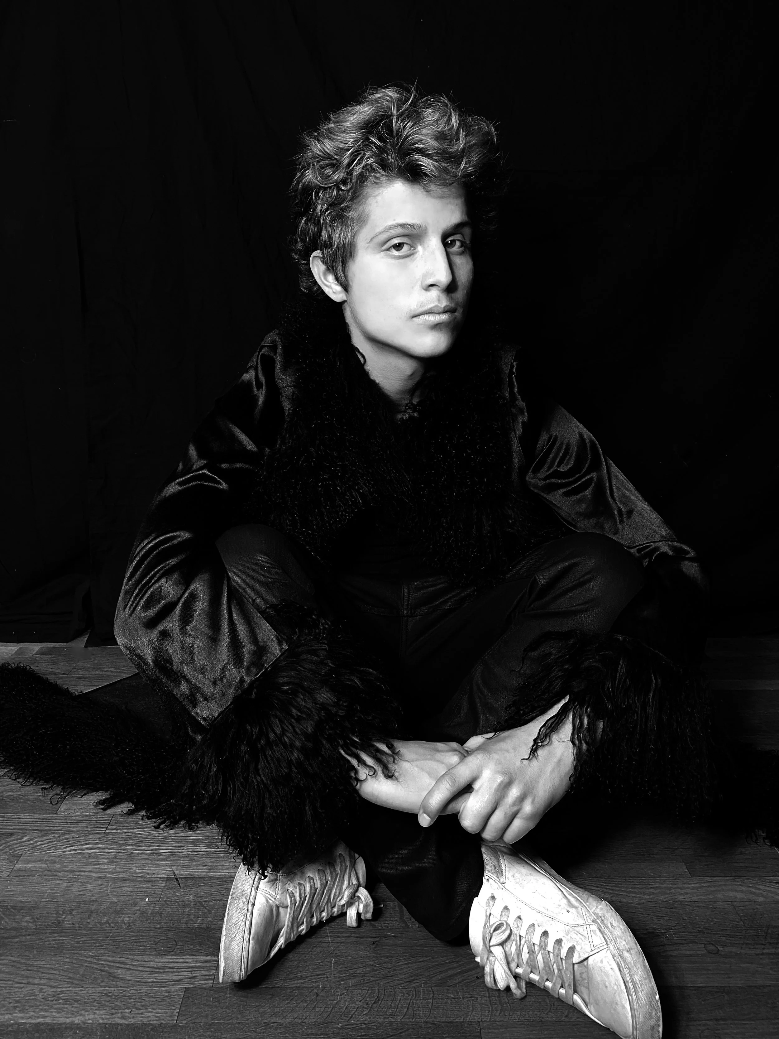 Vancouver BC professional headshot photography. Black and white portrait of a young person sitting cross-legged on the floor, wearing sneakers, with a dark jacket with fur trim, against a dark background.
