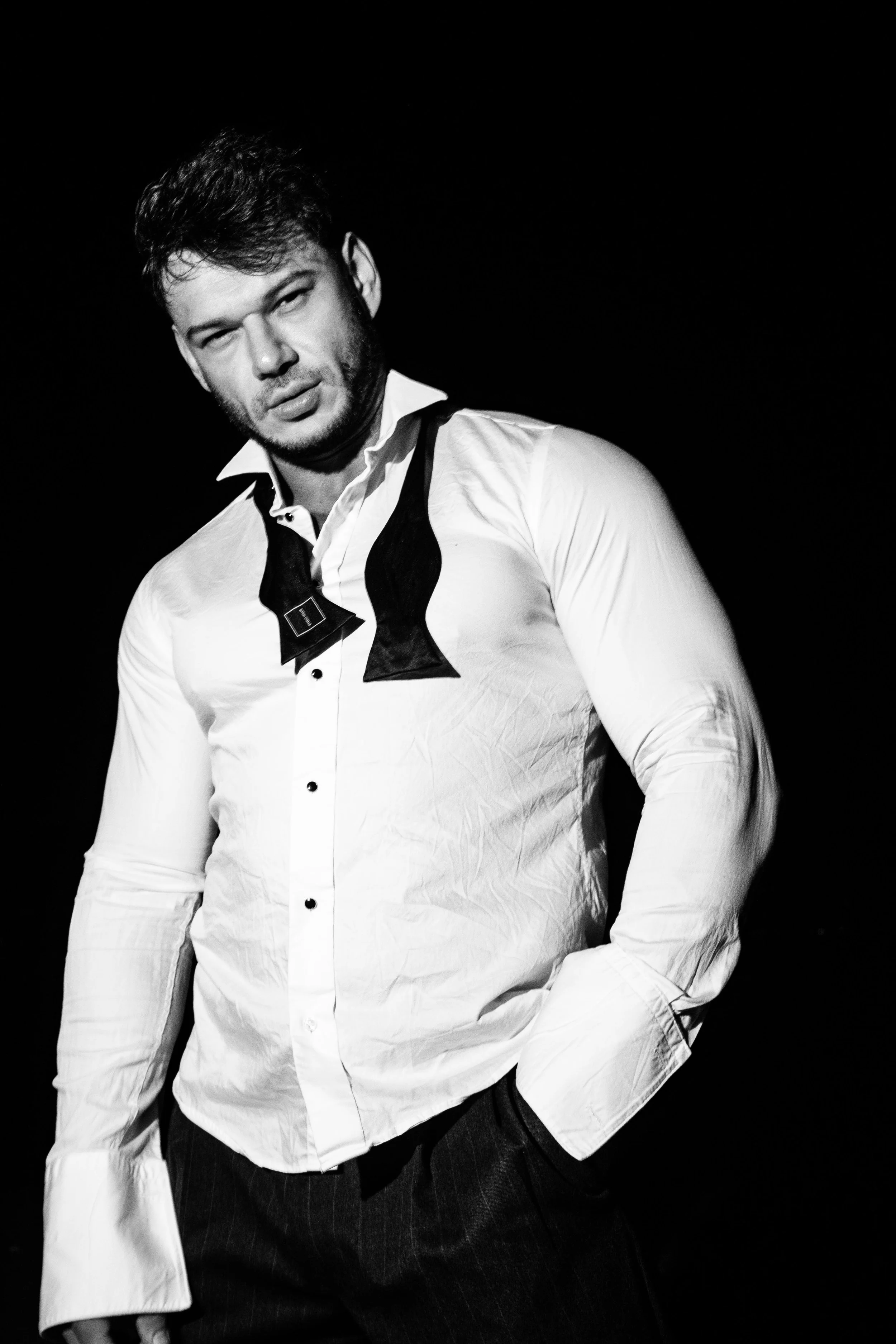 A man in a white shirt with a black bow tie around his neck, standing in front of a black background. The shirt appears to be wrinkled, and he has his hand in his pocket. The image is in black and white.