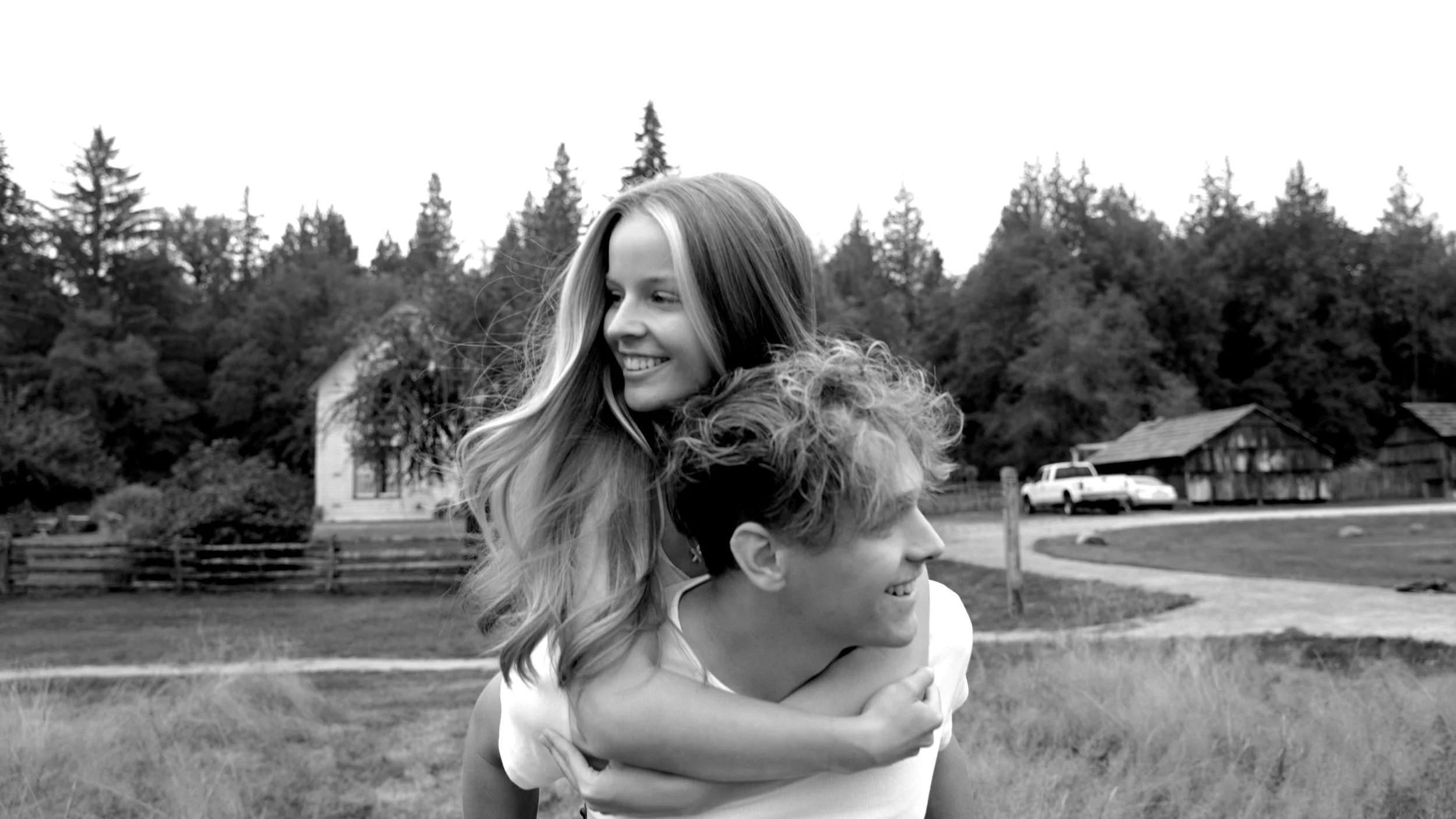 A black and white photo of a girl giving a piggyback ride to a boy in an outdoor rural setting with trees, houses, a car, and a dirt path.Vancouver BC professional headshot photography.