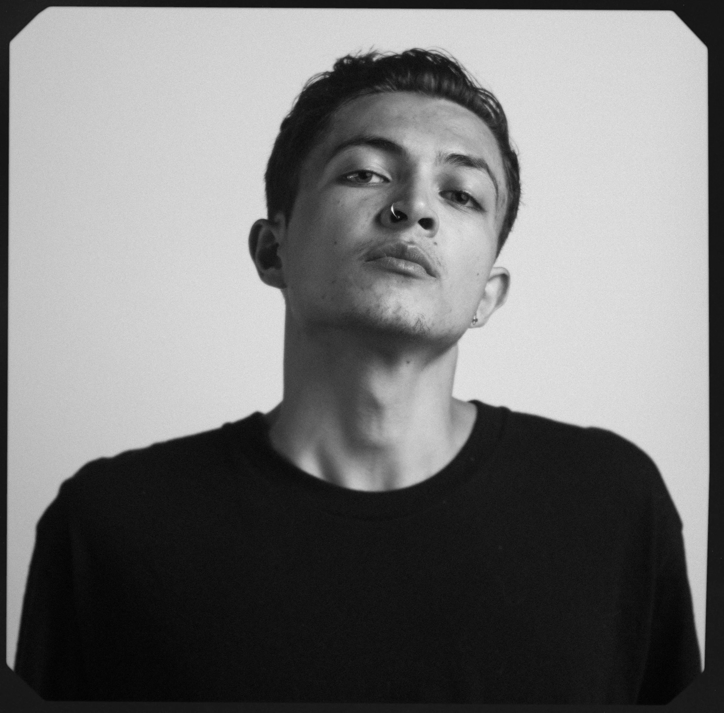 Vancouver BC professional headshot photography. Black and white portrait of a young person with short dark hair, a nose piercing, and earrings, wearing a plain black shirt, looking confidently at the camera against a plain light background.
