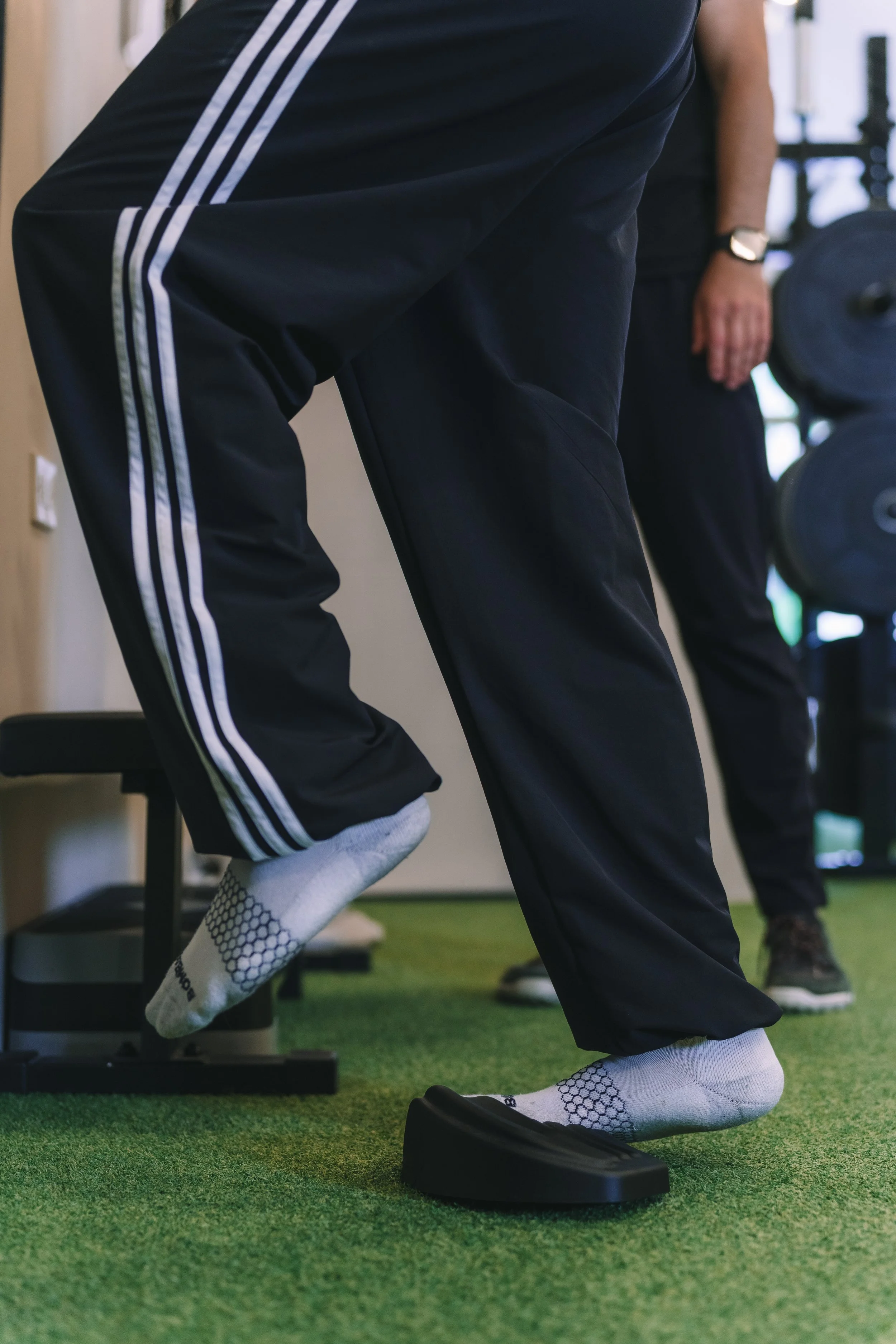 A person performing a squat exercise on a black rocker board with other gym equipment visible in the background, wearing black athletic pants, white honeycomb-patterned socks, and a black watch.