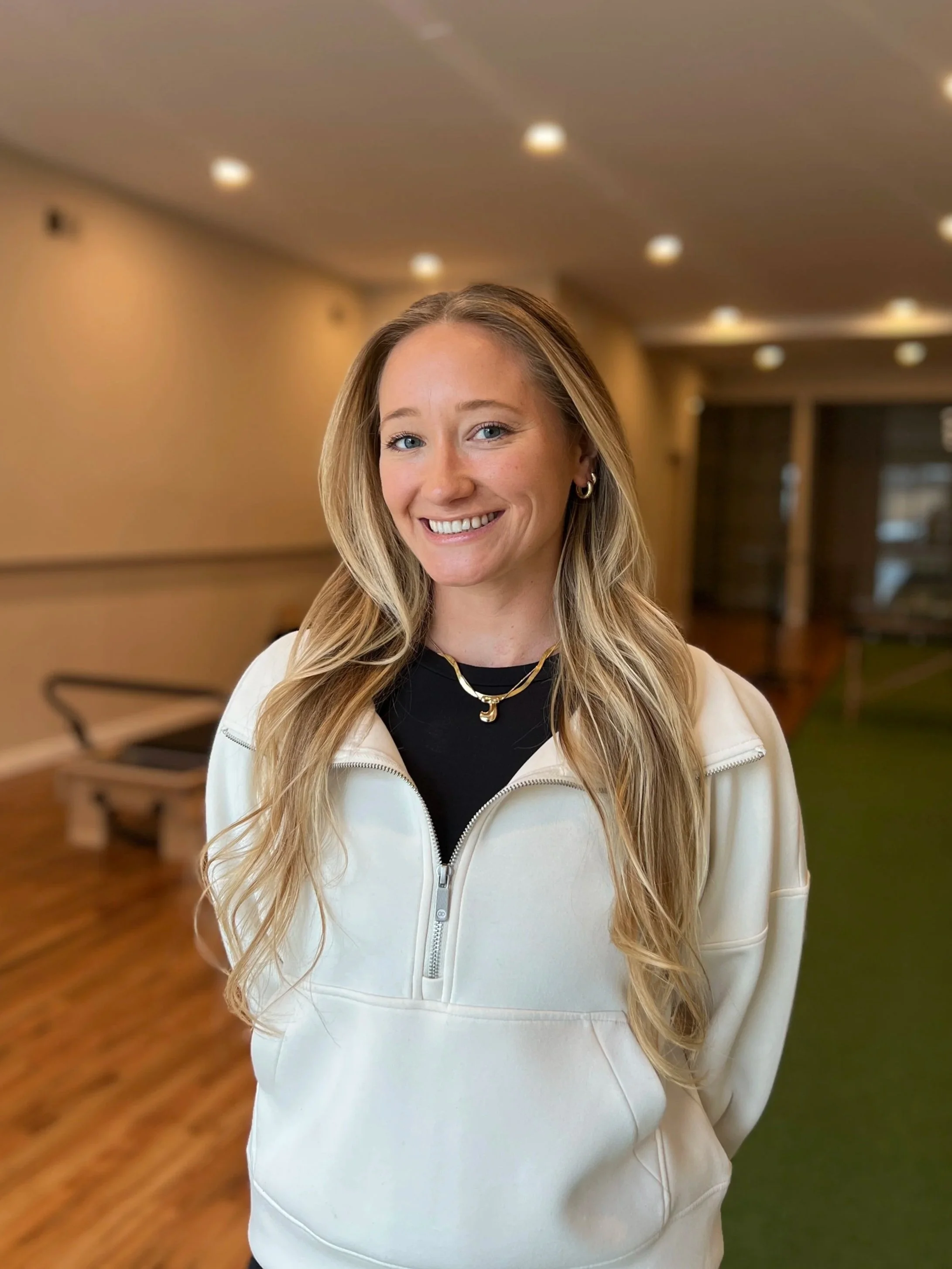 A woman with long blonde hair, smiling, wearing hoop earrings, a black top, a gold chain with a pendant, and a white zip-up jacket, standing in an indoor space with brown walls, wooden flooring, and ceiling lights.