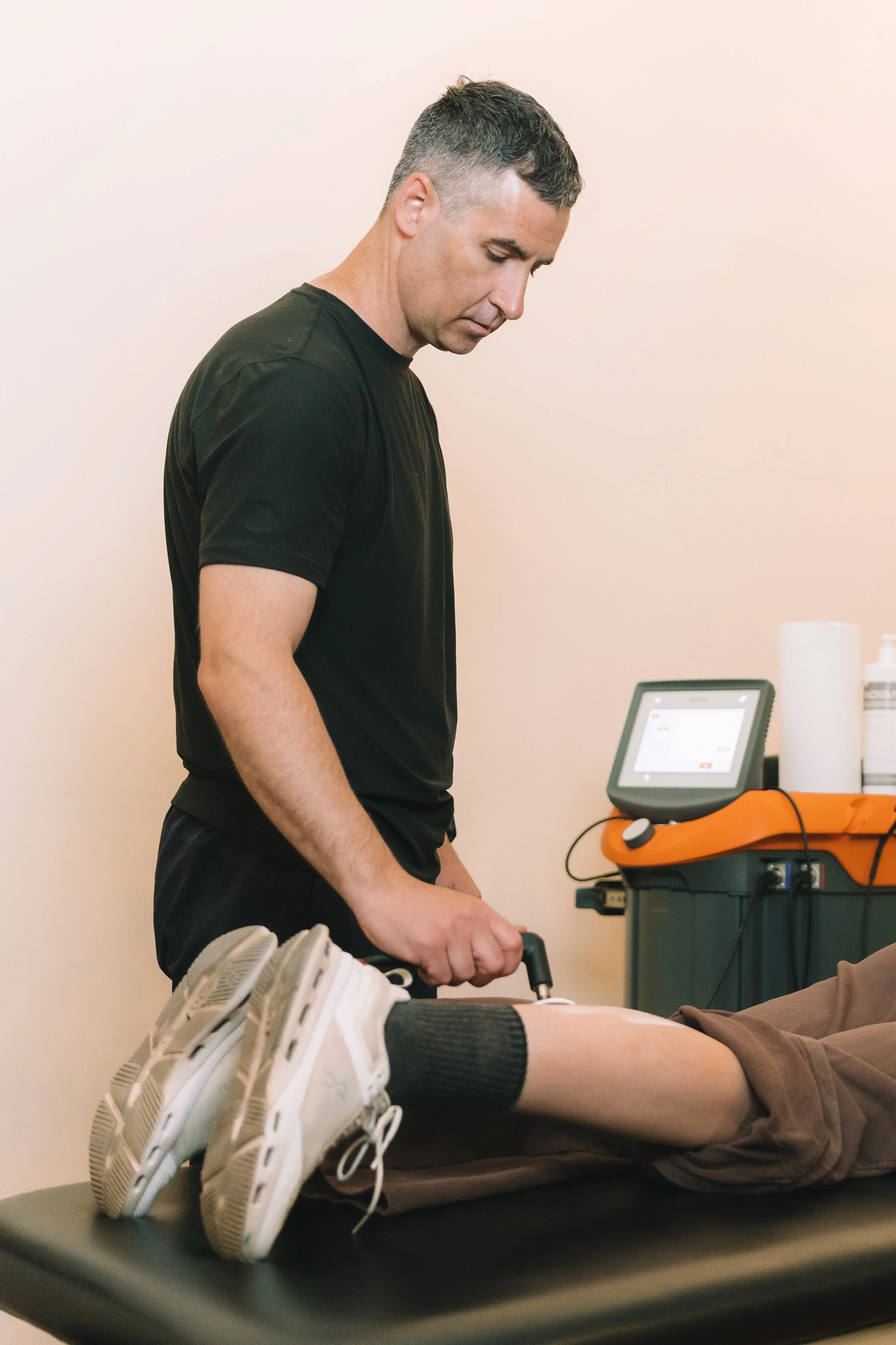 A man in black shirt and dark pants preparing to give an ultrasound to a patient lying on an examination table, with their legs and shoes visible in the foreground.