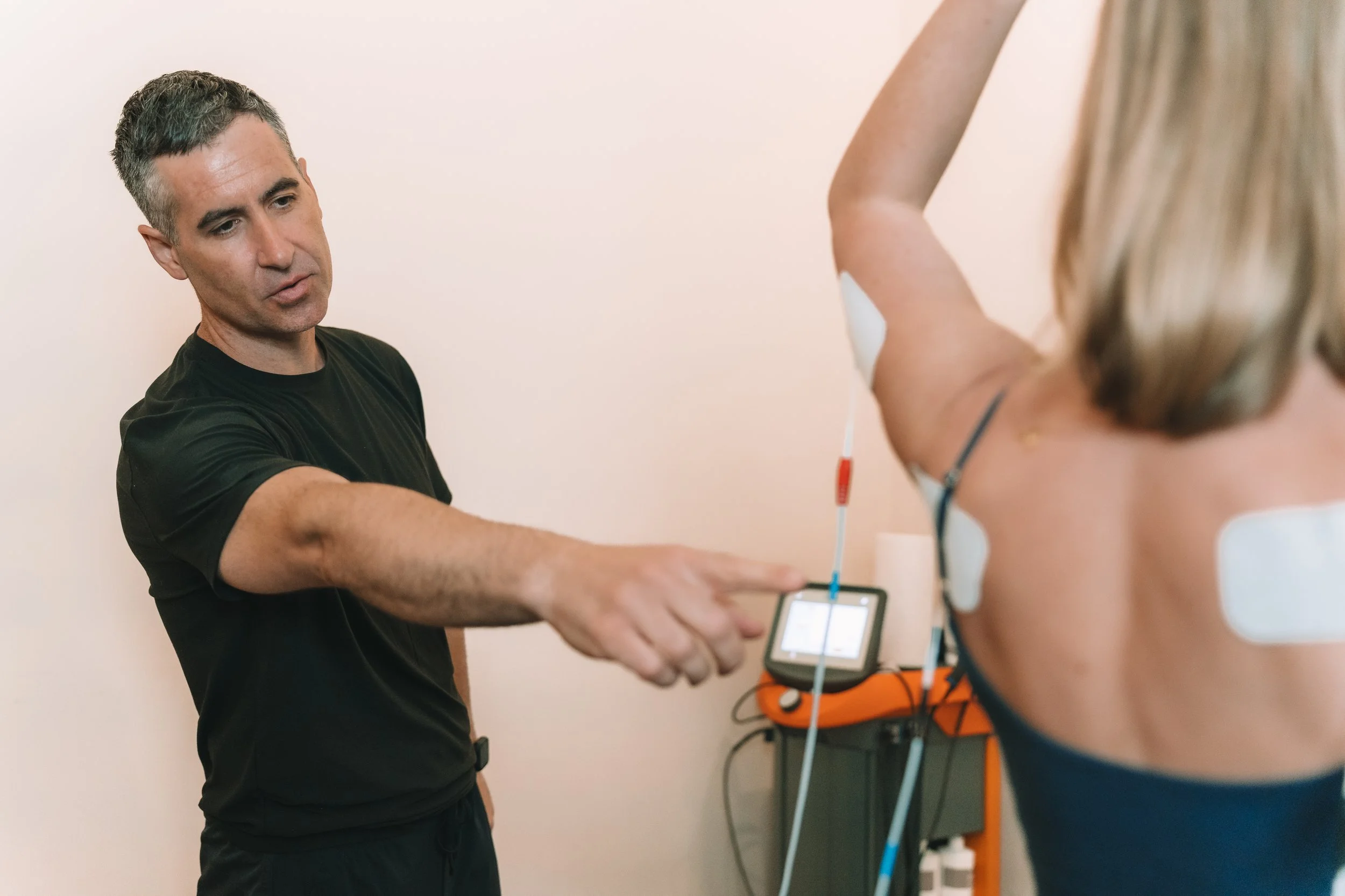 A male healthcare professional performing a medical test on a woman who has medical electrodes attached to her chest, with medical equipment and monitors visible in the background.