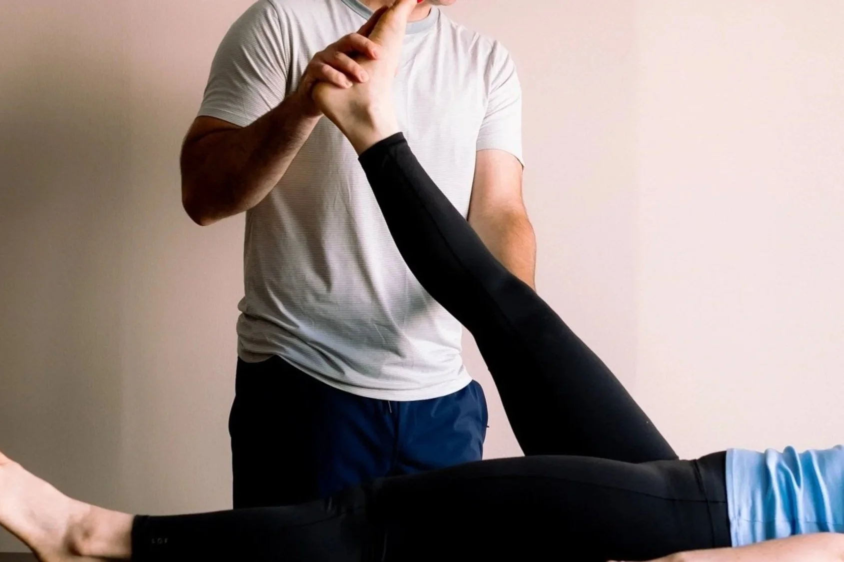 A person lying on her stomach receiving a physical therapy or chiropractic adjustment from a therapist standing beside her. The patient is wearing black leggings and light blue socks, while the therapist is in a white T-shirt and dark shorts, holding the patient's arm and back.