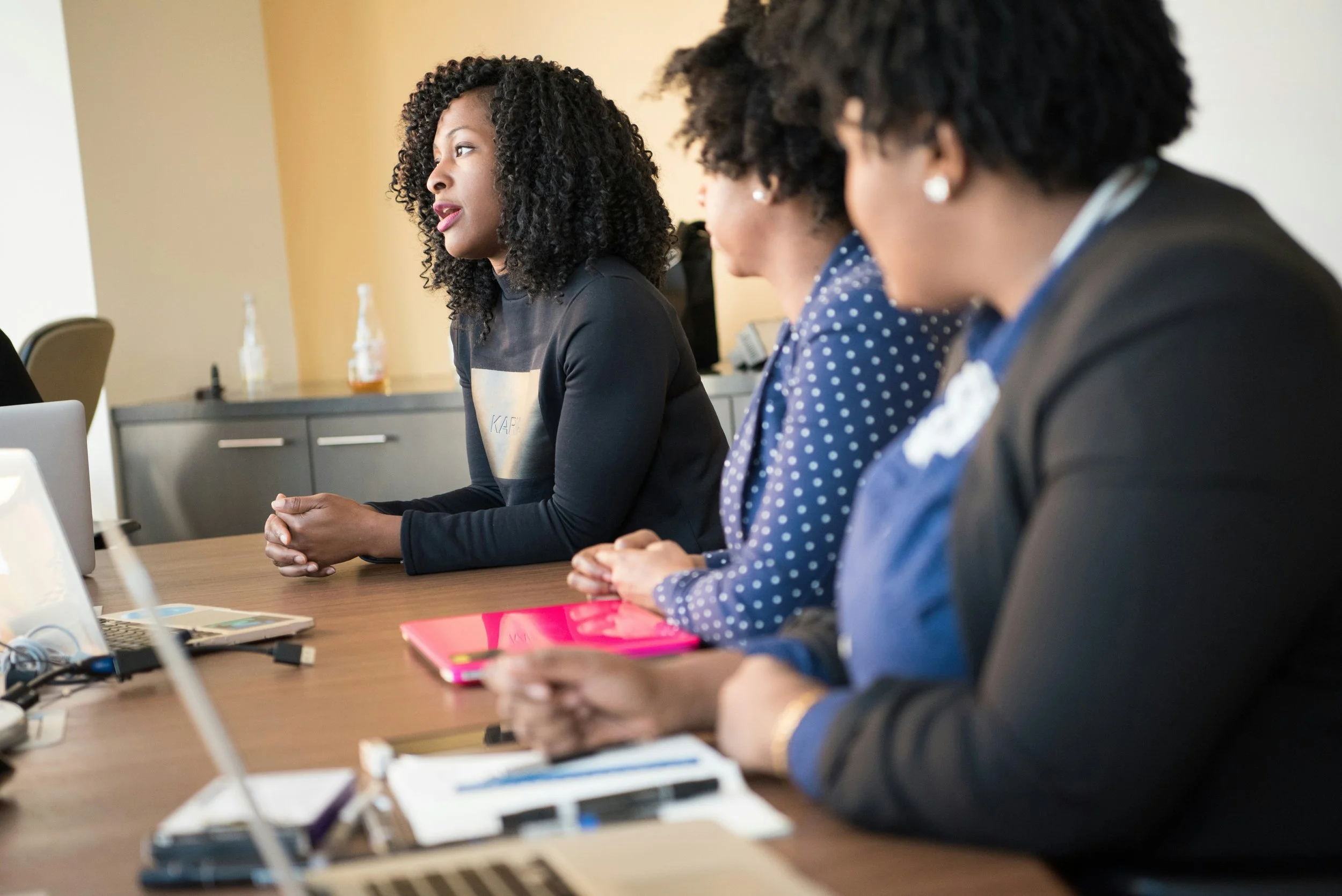 Four women sitting at a conference table, listening to a presentation or discussion in a professional setting.