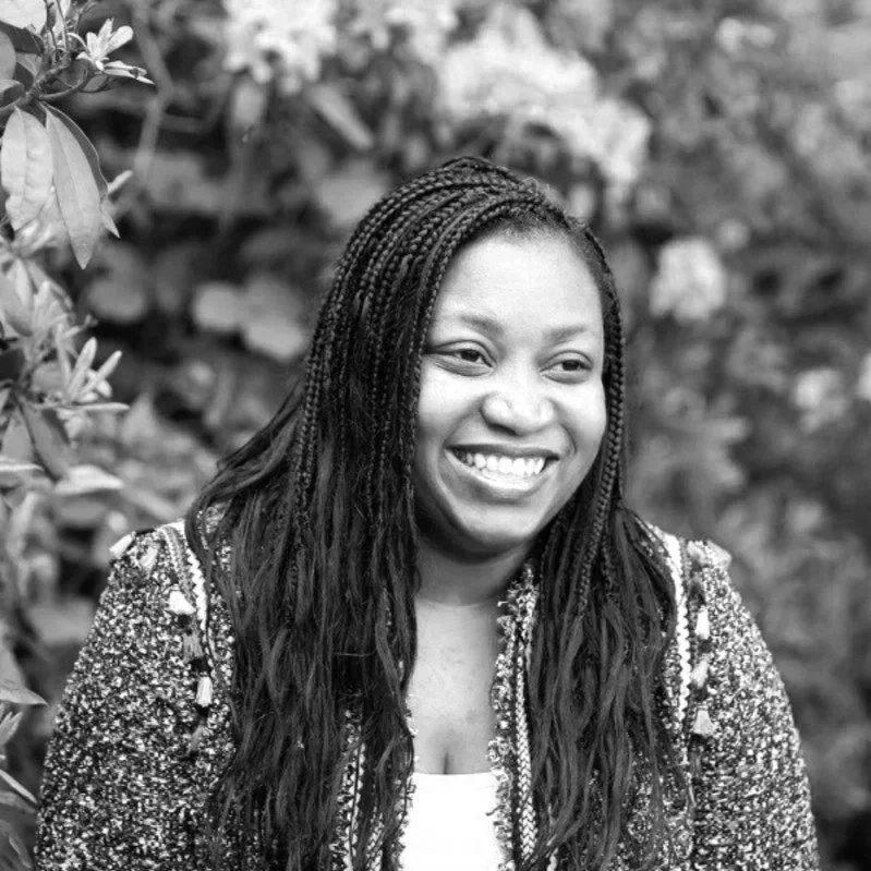 A smiling woman with long braided hair, wearing a patterned jacket outdoors with leafy background.