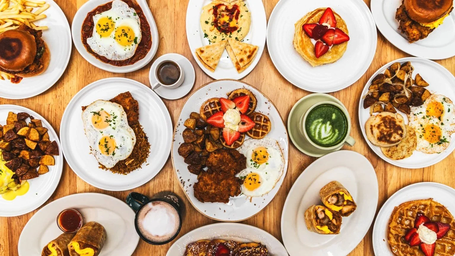 A variety of breakfast dishes on a wooden table, including eggs, pancakes, waffles, hash browns, breakfast wraps, and beverages.