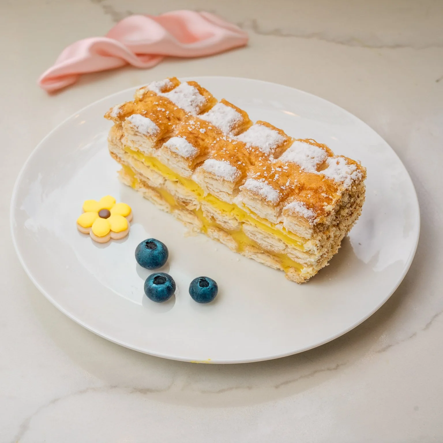 A slice of layered cream-filled pastry cake dusted with powdered sugar on a white plate, garnished with three blueberries and a small yellow flower-shaped candy. A pink cloth is in the background on a white marble surface.
