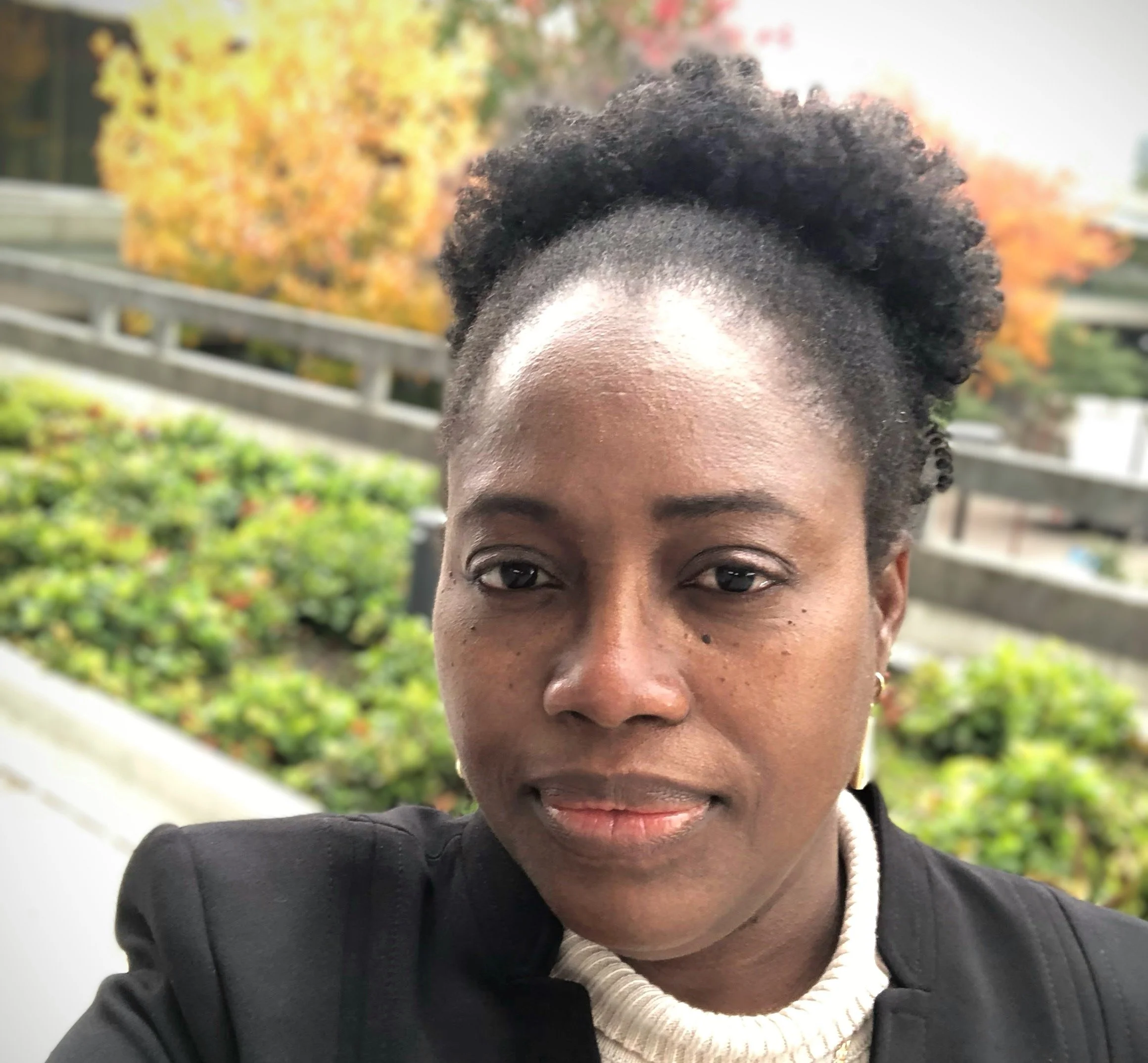 A close-up of a woman with dark skin and natural curly hair styled up, wearing a white knit turtleneck and a black jacket, outdoors with autumn-colored trees and greenery in the background.
