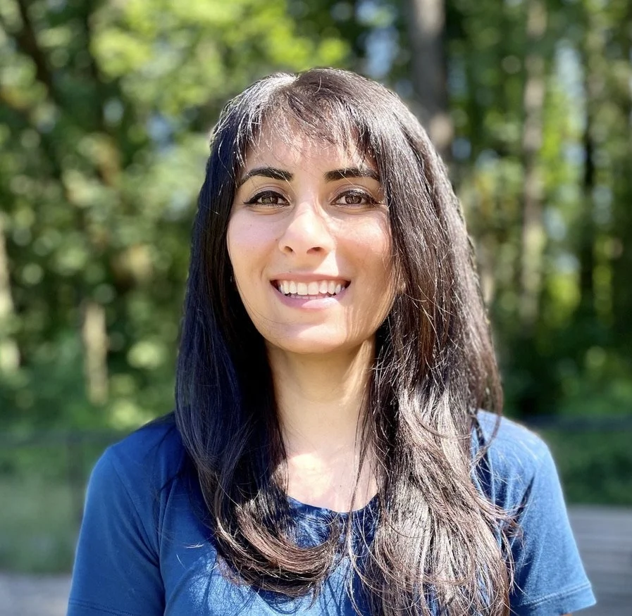 A smiling woman with dark hair outdoors in a sunny, green, wooded area.