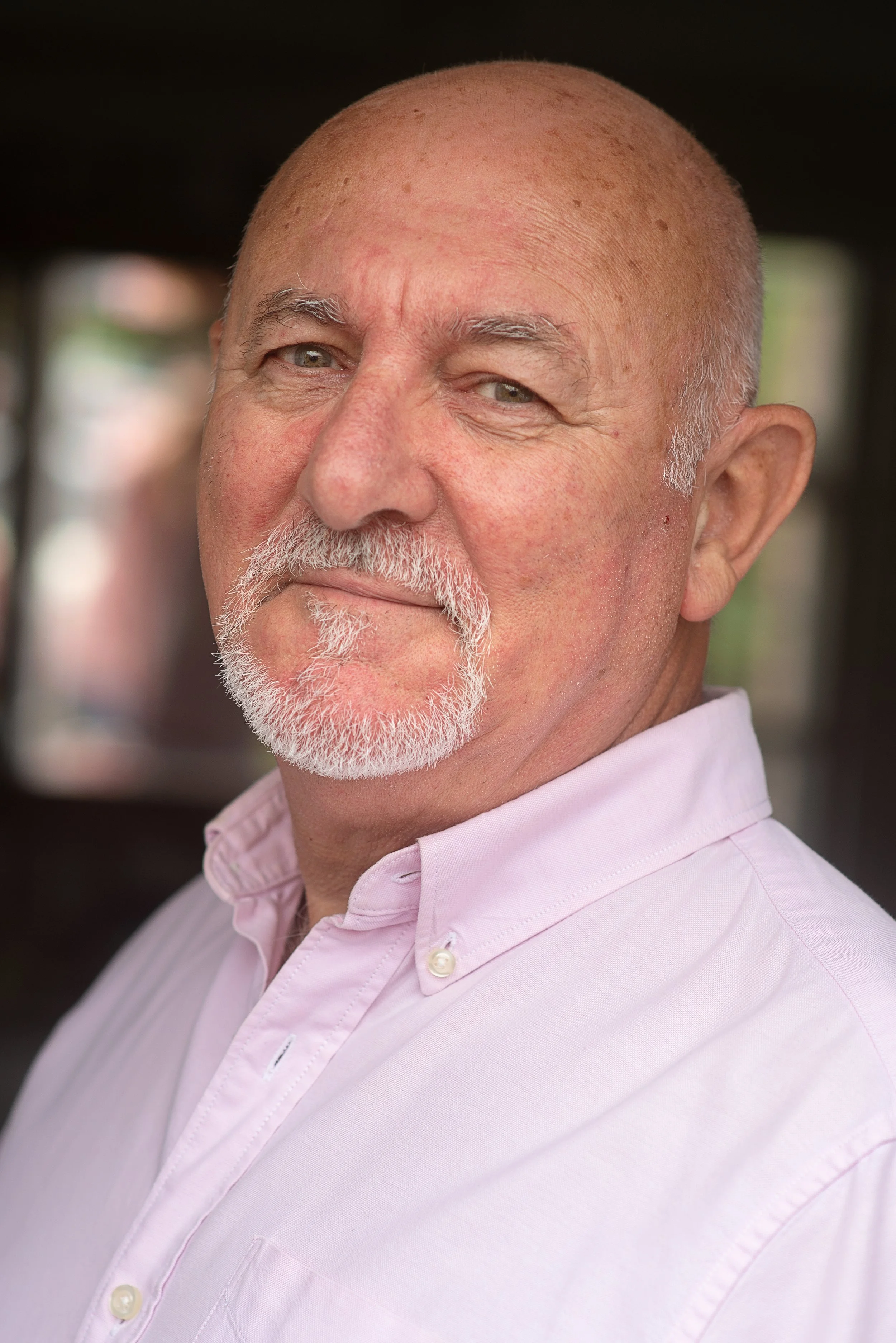 Close-up portrait of an older bald man with a white beard and mustache, wearing a light pink collared shirt, smiling slightly.