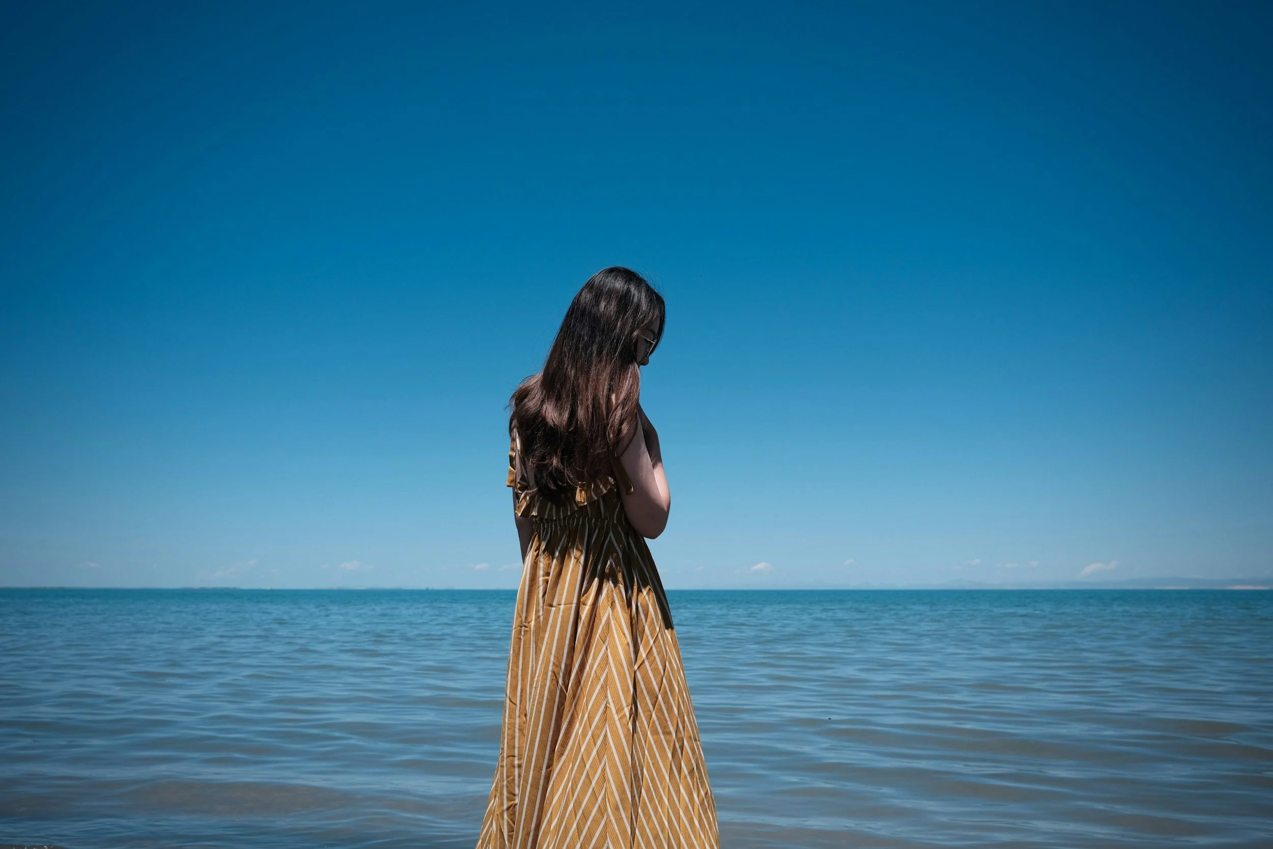 A woman with long dark hair wearing a yellow and brown striped dress stands in shallow water at the beach, looking down with her arms crossed, against a clear blue sky and distant horizon.