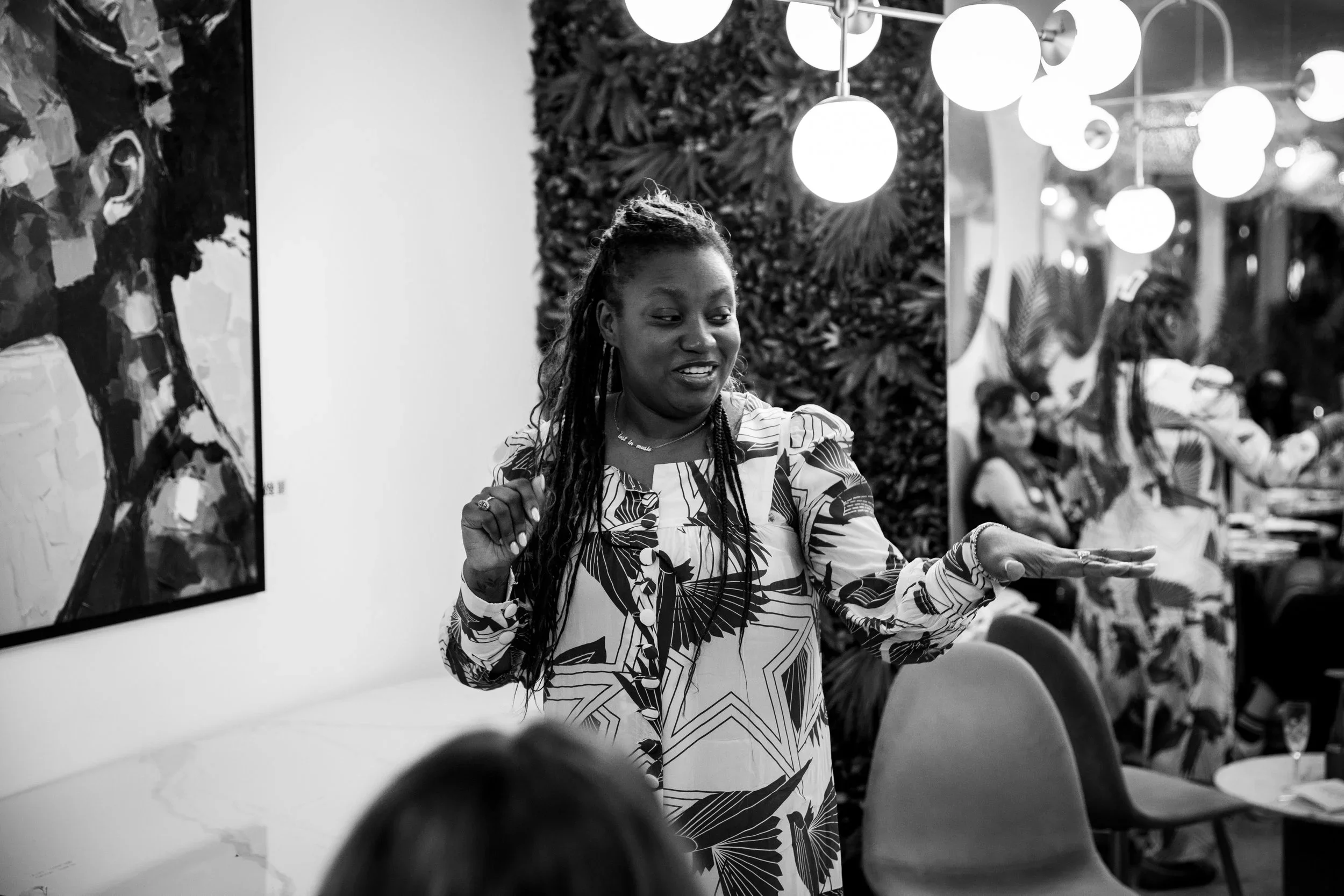A woman with long dreadlocks, wearing a patterned dress, is speaking or gesturing with her hands in an indoor setting. She is standing next to a wall with a painting and an art piece with plants. Several people are seated and listening in the background.