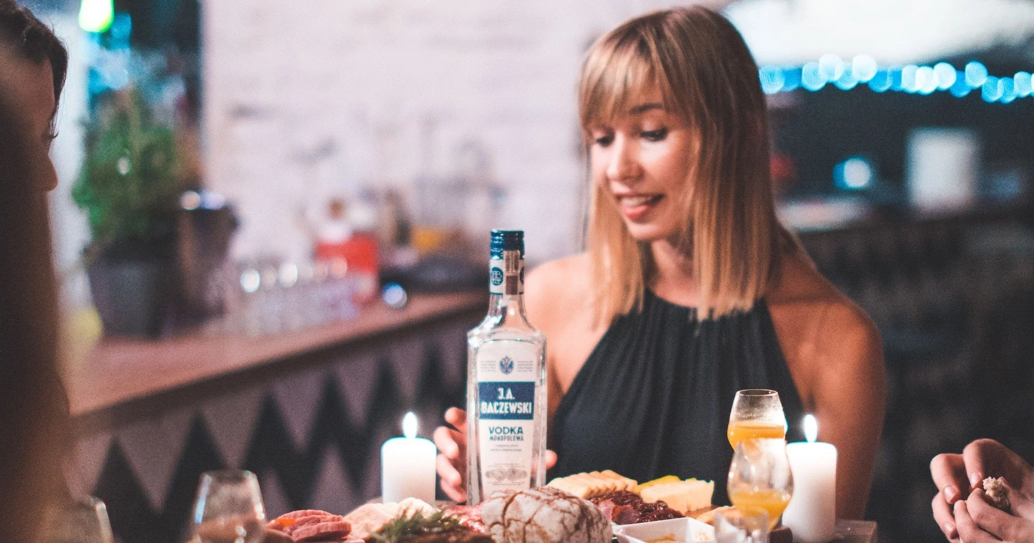 A woman with shoulder-length blonde hair wearing a black sleeveless top is sitting at a party table with food and drinks, holding a bottle of vodka. She is smiling and engaging with others, with candles and glasses on the table, in a cozy, dimly-lit setting.
