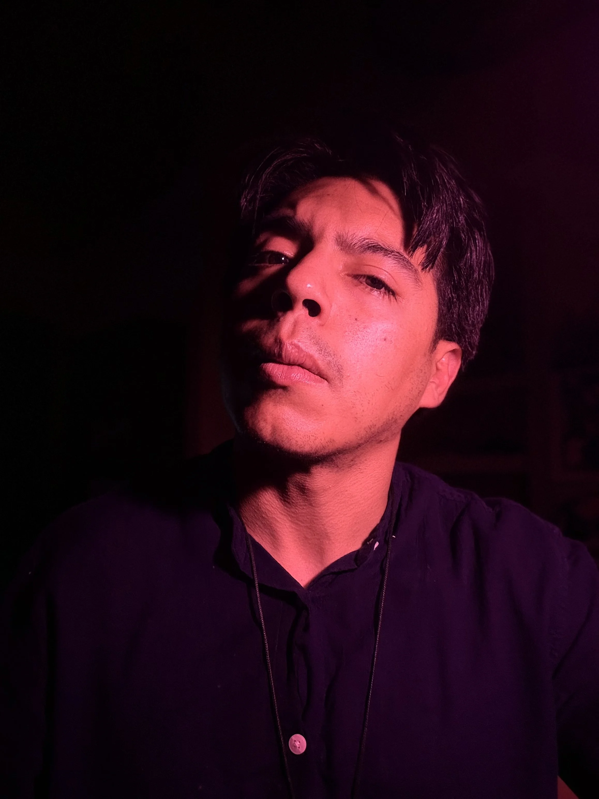 A young man with dark hair and a light beard, bathed in pink and purple lighting, wearing a black shirt with a necklace, looking directly at the camera.