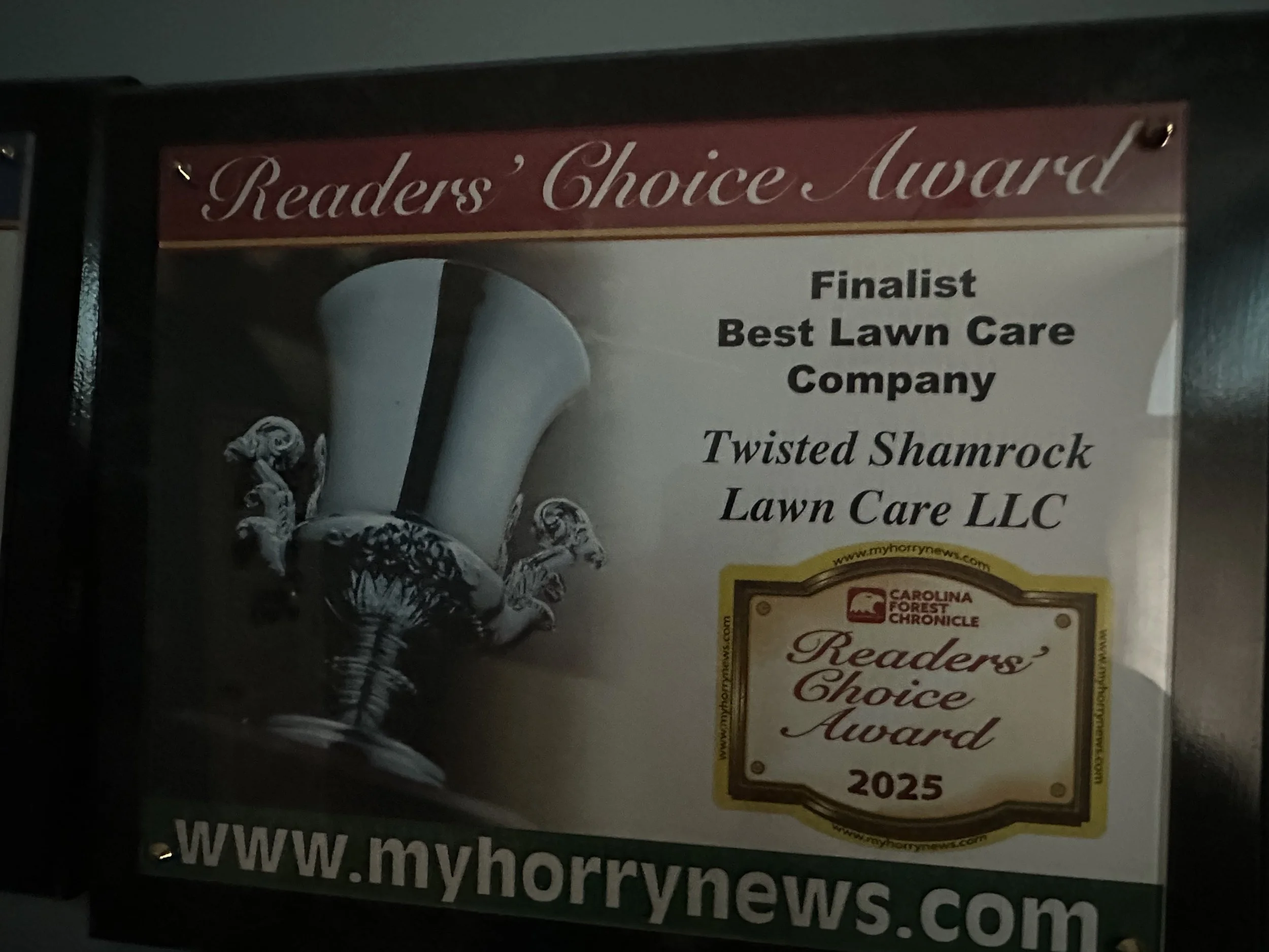 A sign showing a Readers' Choice Award for Twisted Shamrock Lawn Care LLC, finalist for Best Lawn Care Company, with a decorative trophy and a yellow border around the award details. The website is www.myhorrynews.com.