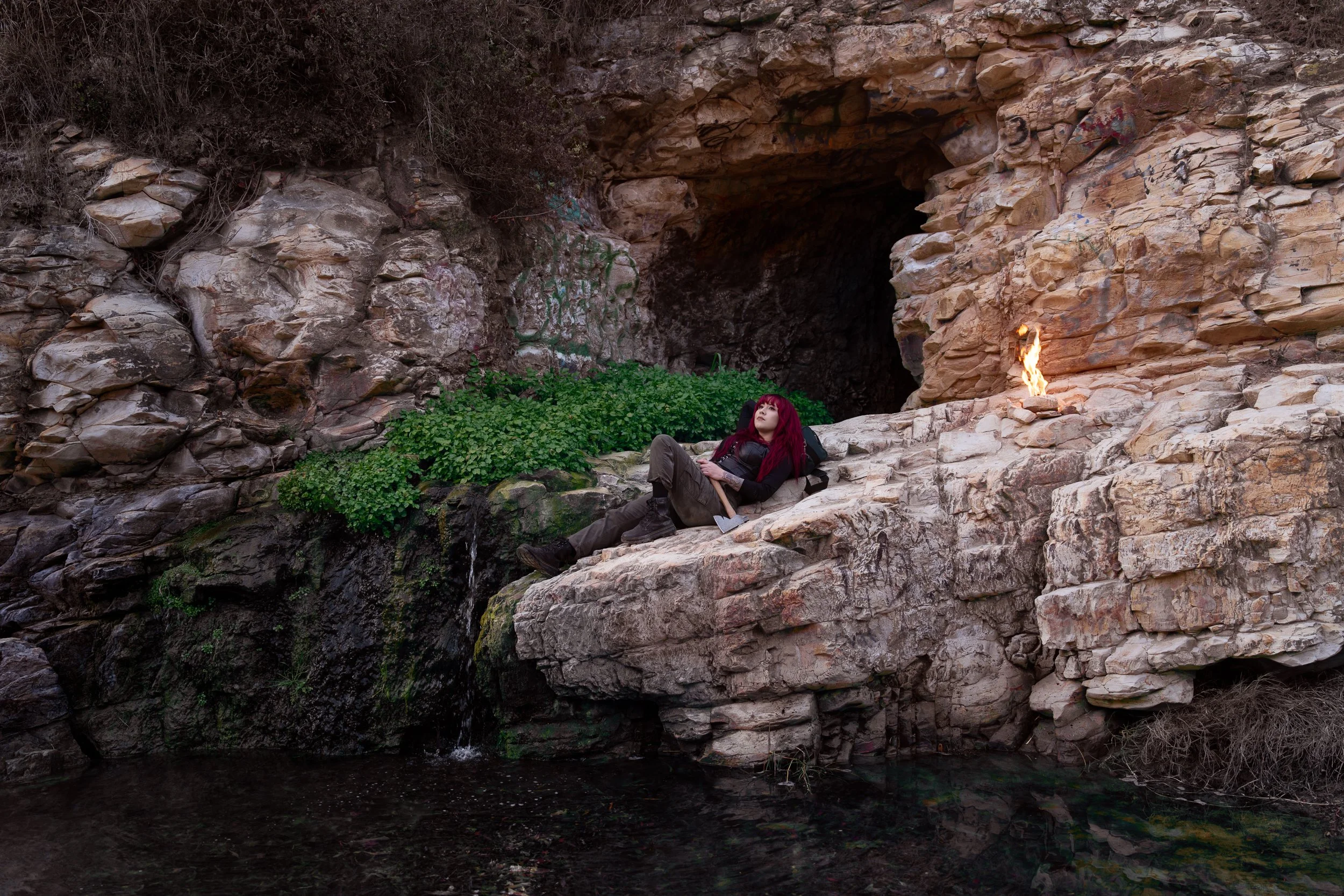 A woman with red hair and dark clothing lying on a rock ledge in front of a cave, with a small fire burning nearby and water flowing beneath her.