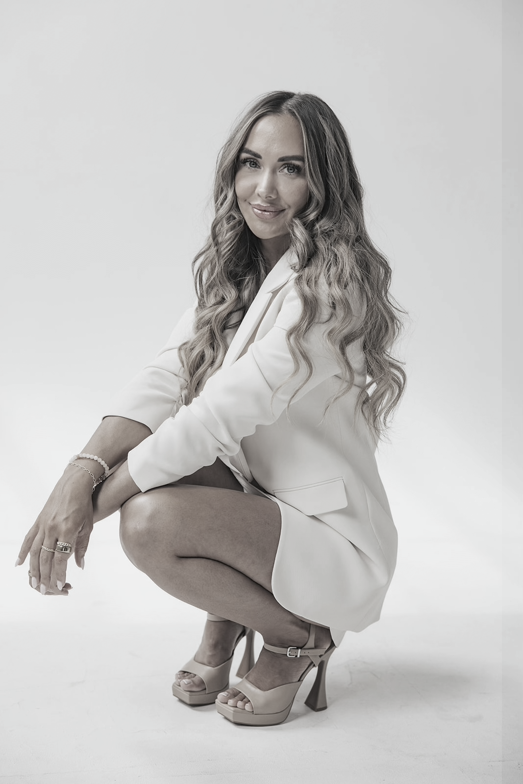 A woman with long wavy hair in a white blazer and skirt, wearing high heels, squatting against a plain light background.