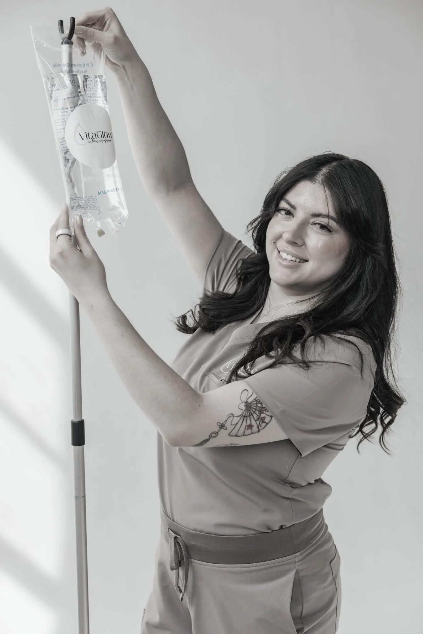 A woman with dark hair and tattoos on her arm, smiling while holding a clear IV bag labeled Vitaglow. She is wearing a scrub top and pants, standing against a plain light background.