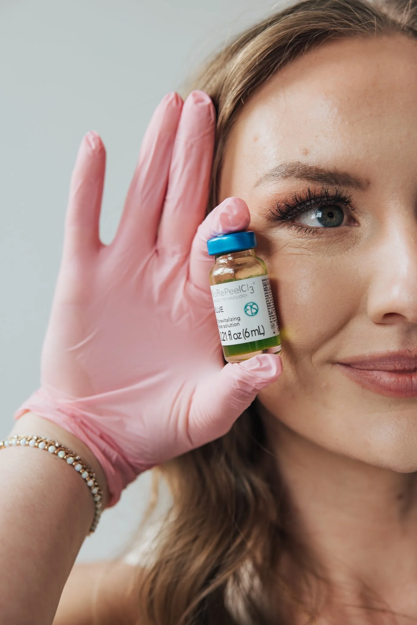 A woman smiling while holding a small vial labeled 'BioRePeelCl3' close to her face, wearing a pink glove on her hand.
