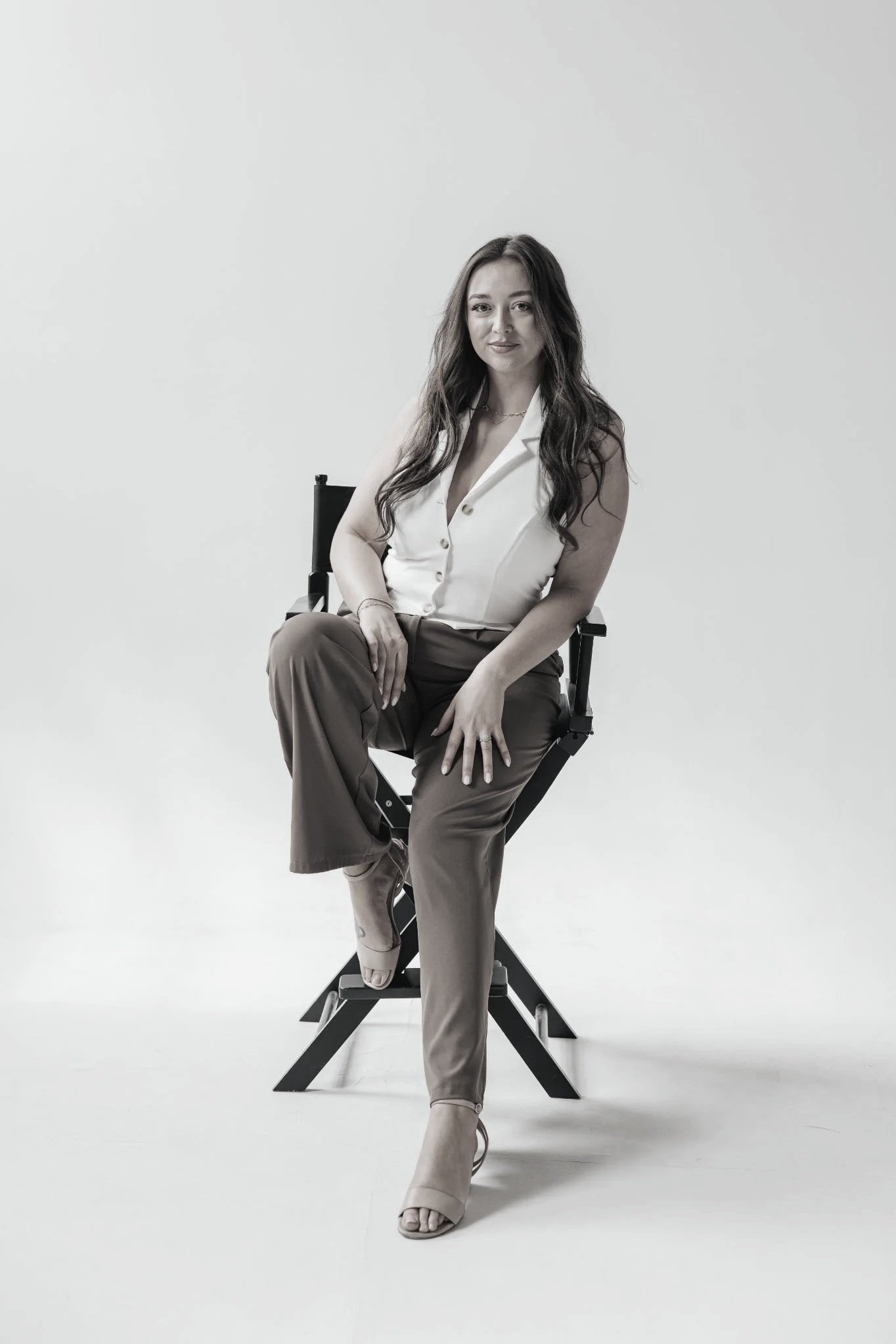 A woman with long wavy hair sitting on a director's chair in a minimalist setting, dressed in a sleeveless white blouse and brown trousers.