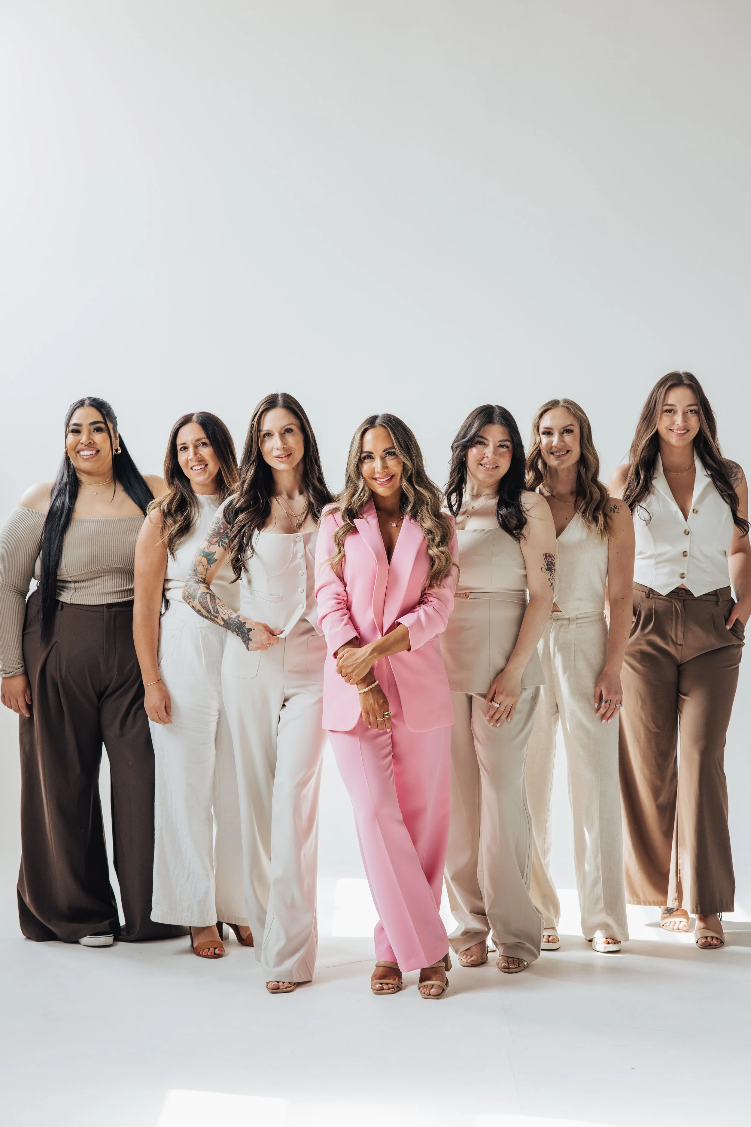 Group of eight women standing together in fashionable outfits, posing for a photo with a plain white background.
