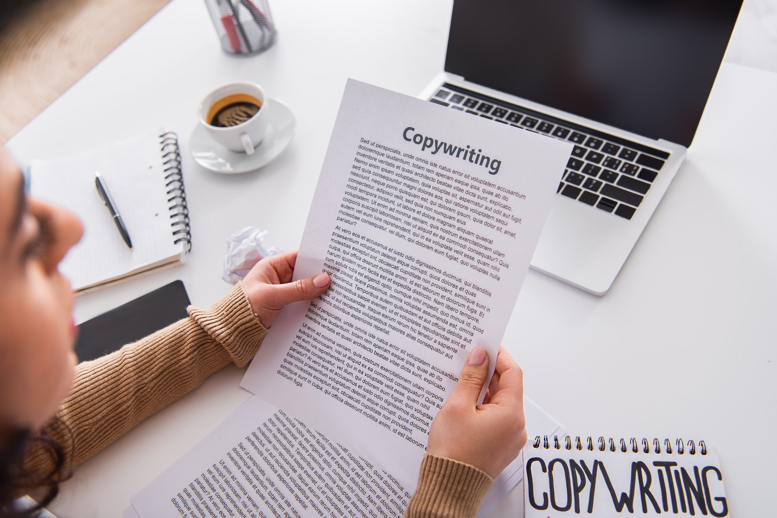 Person holding a printed copywriting document at desk with a laptop, cup of coffee, notepad, and pen nearby.